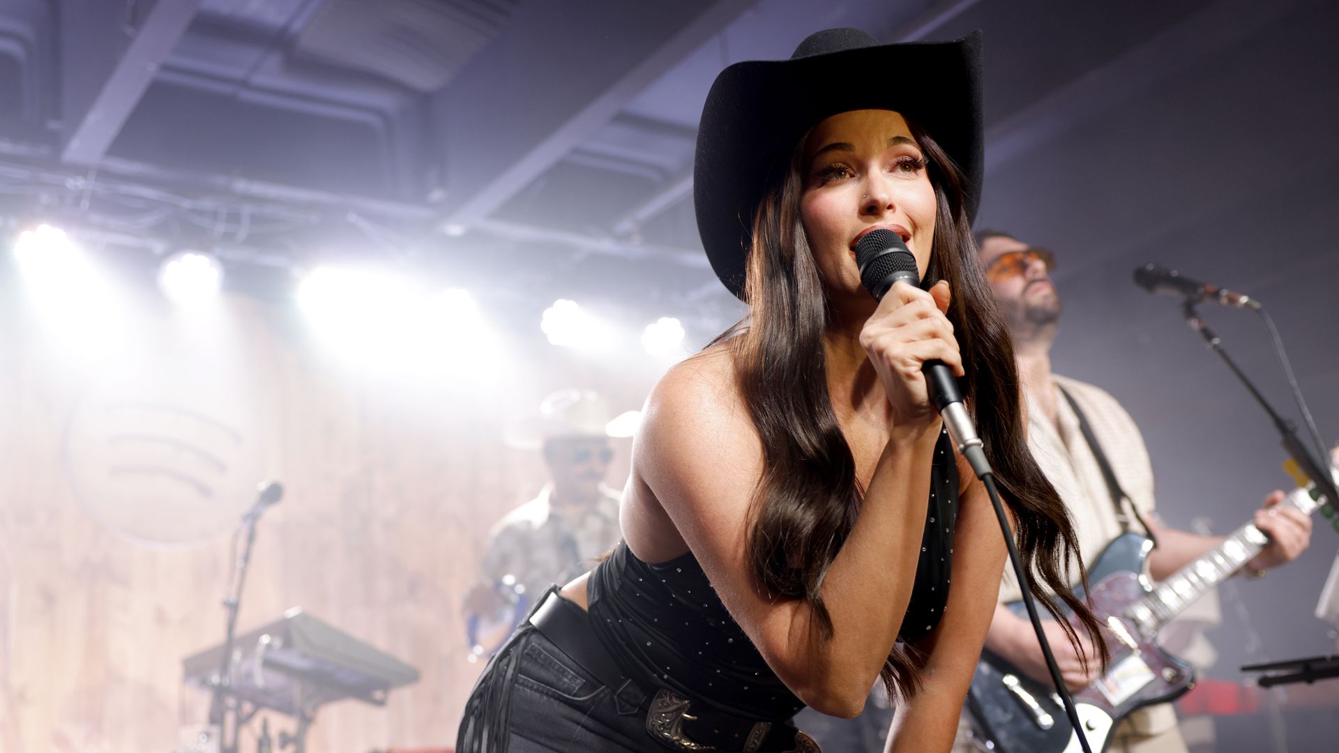 A female singer wearing a black cowboy hat sings into a microphone on a bright stage, with a guitarist and stage gear blurred in the background.