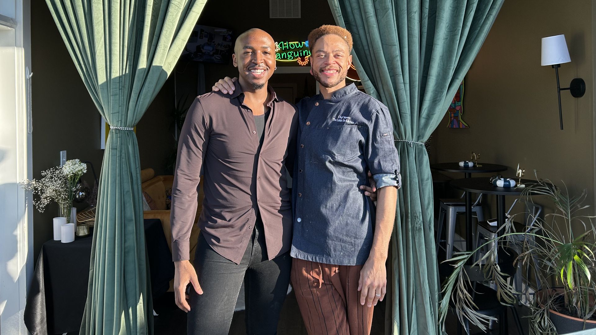 Two smiling men standing arm in arm between teal curtains. One wears a dark shirt and black pants, the other a chef's coat and striped pants. Indoor setting with plants and a lamp.