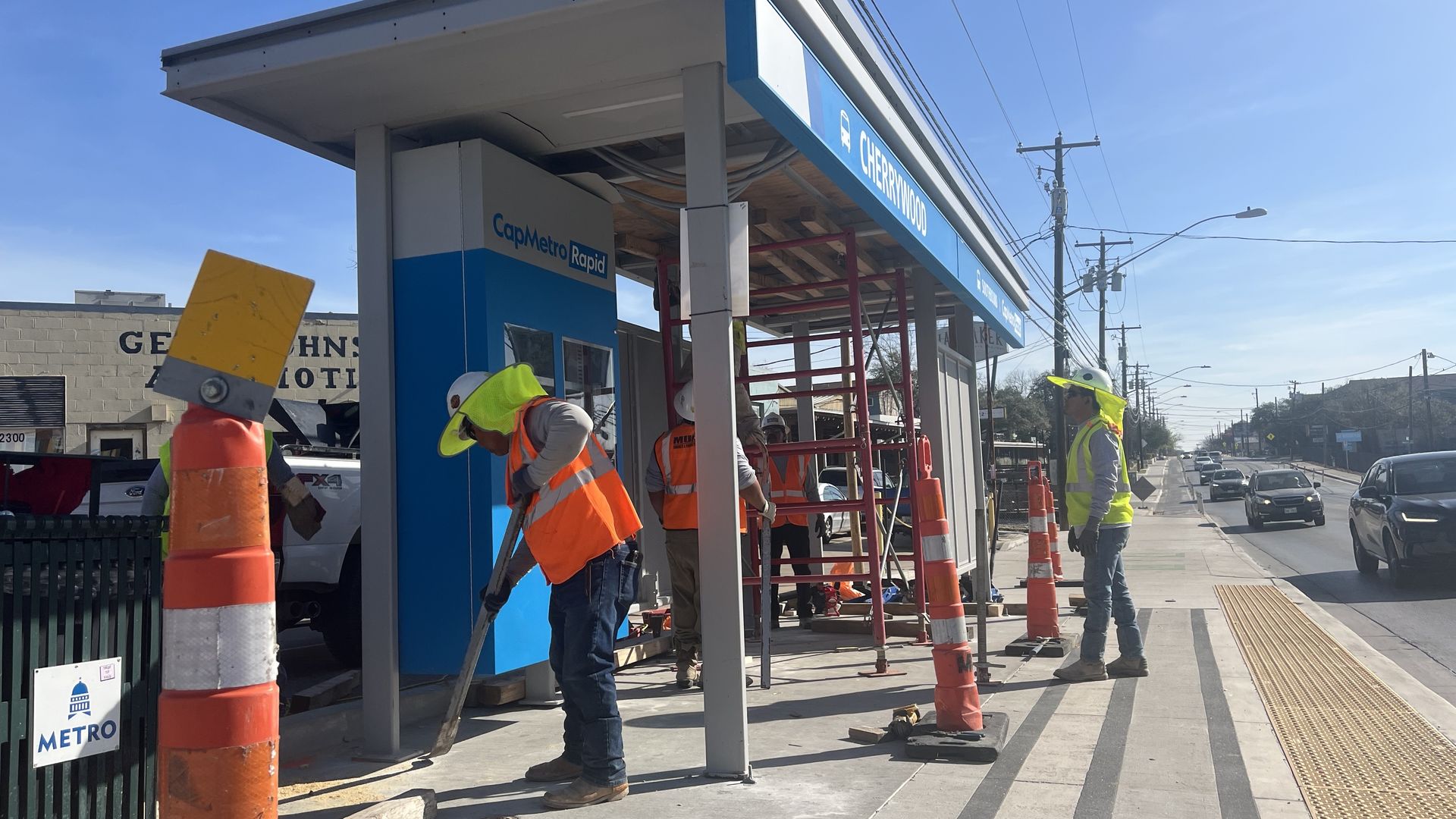 Construction workers wearing orange and yellow safety vests and helmets work on building the Cherrywood CapMetro Rapid bus stop shelter with traffic cones nearby on a sunny day.