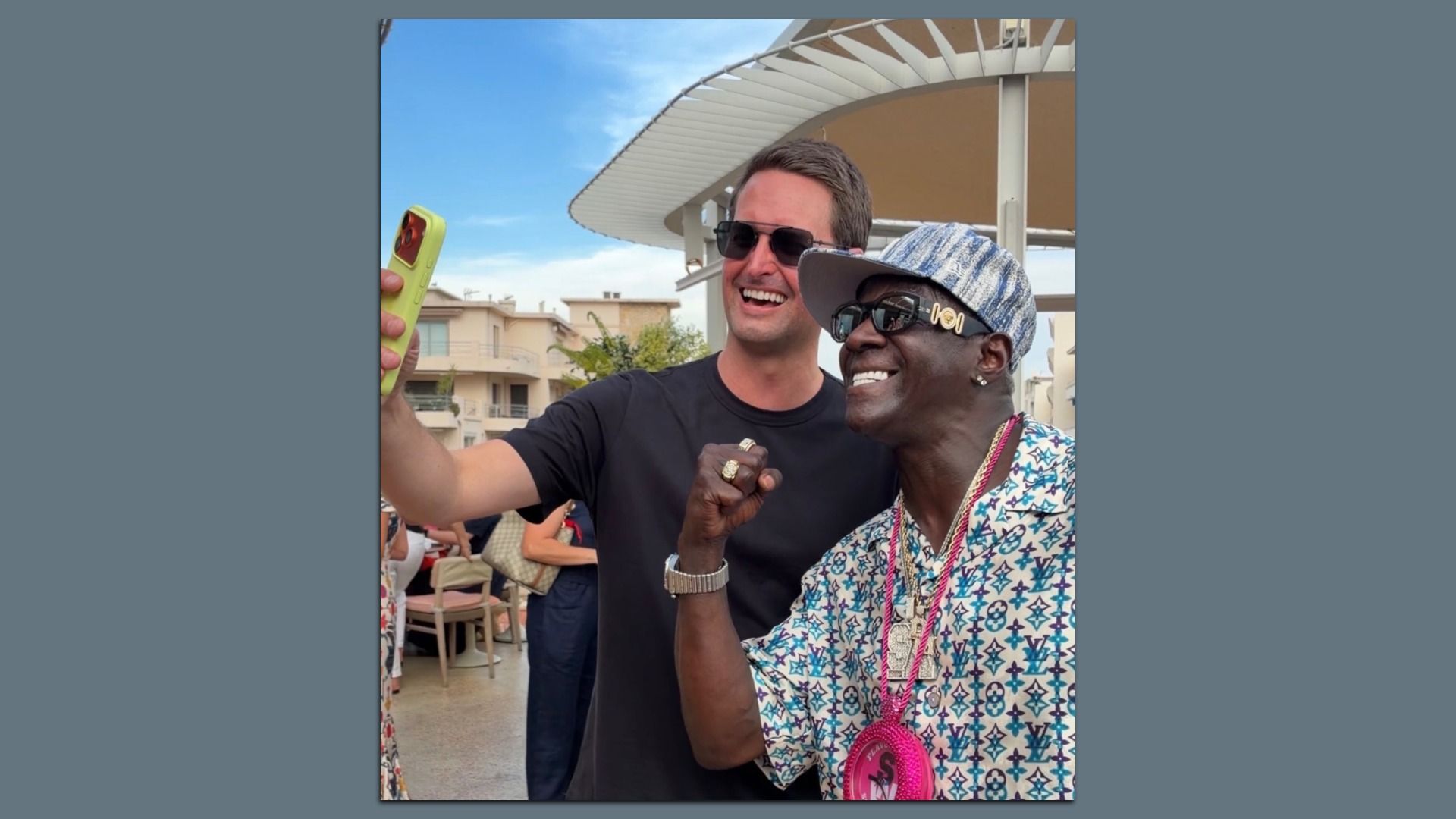 Two men take a sunny selfie outdoors. One wears a black T-shirt and sunglasses, the other in a patterned shirt and striped hat with jewelry. The man on the left holds a bright lime-green phone case.