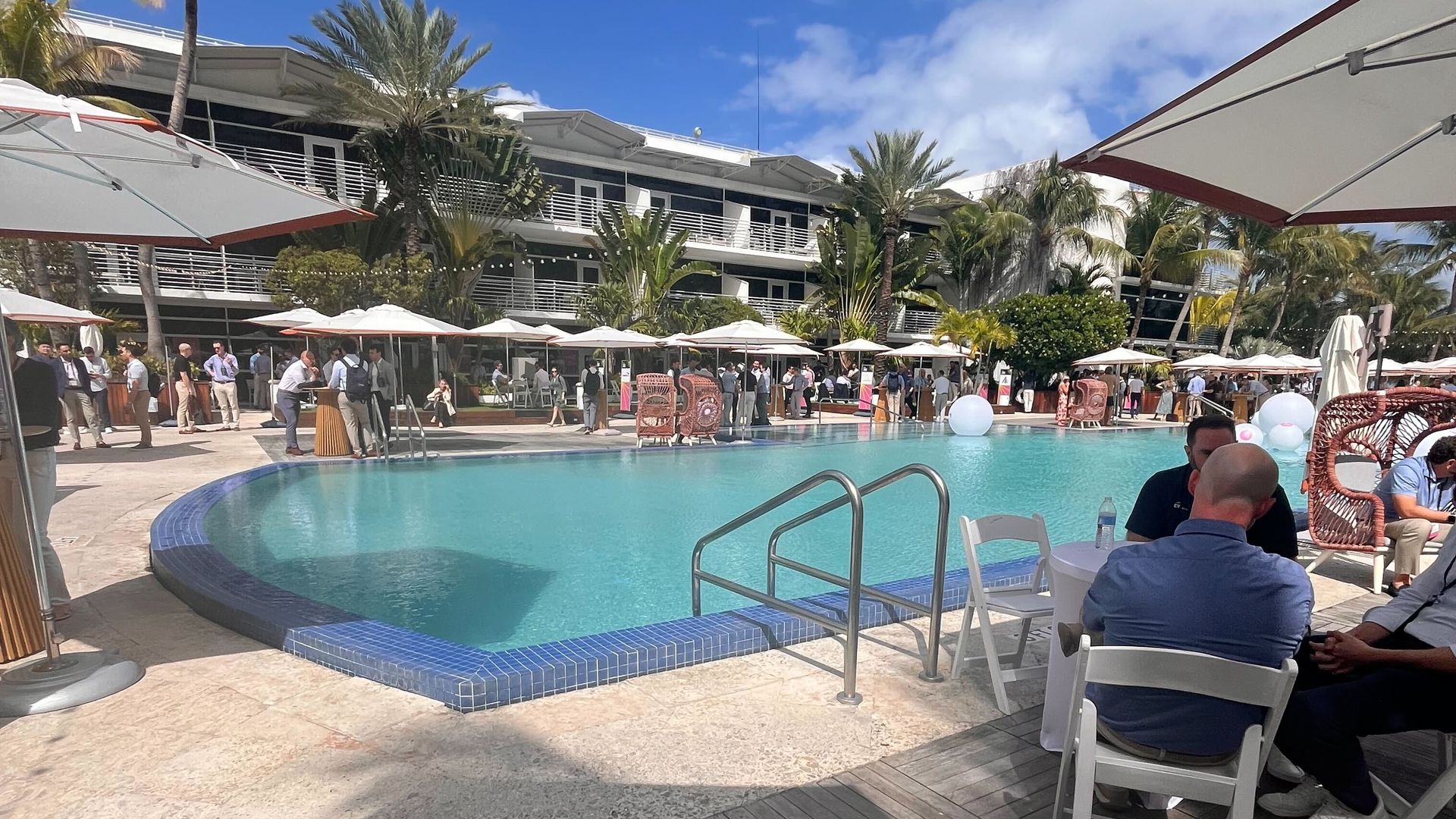 An outdoor pool with several umbrellas and tables around it with people sitting 