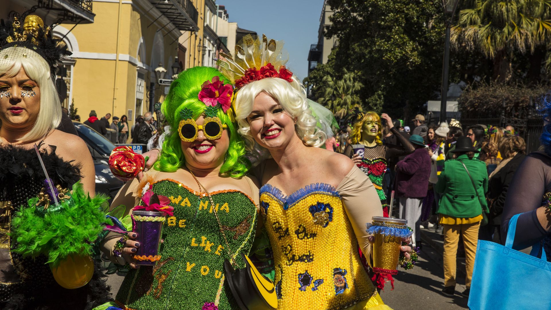 Two Mardi Gras revelers smile as they pose for the camera. They are in costume, wearing embellished corsets and wigs.