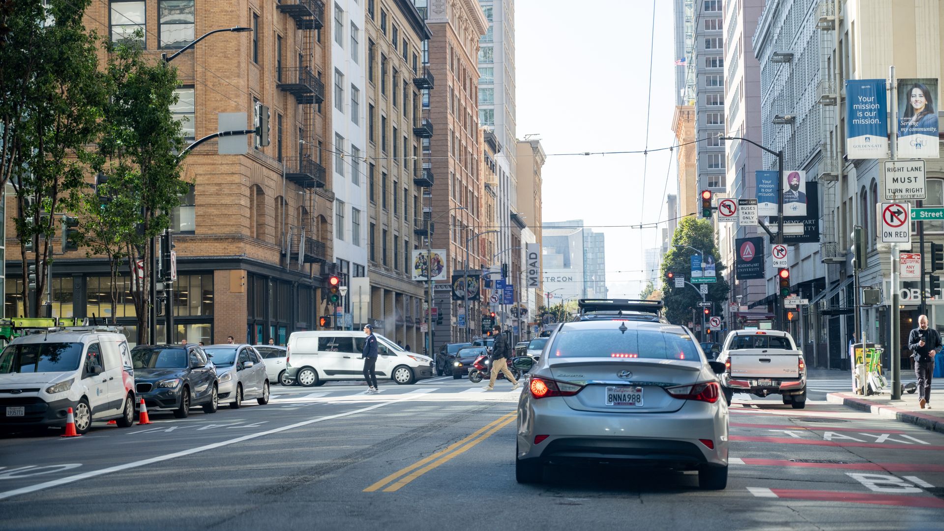 City street scene with cars stopped at red lights, pedestrians crossing, tall beige and gray buildings, green trees, and multiple traffic and mission-themed banners on poles.