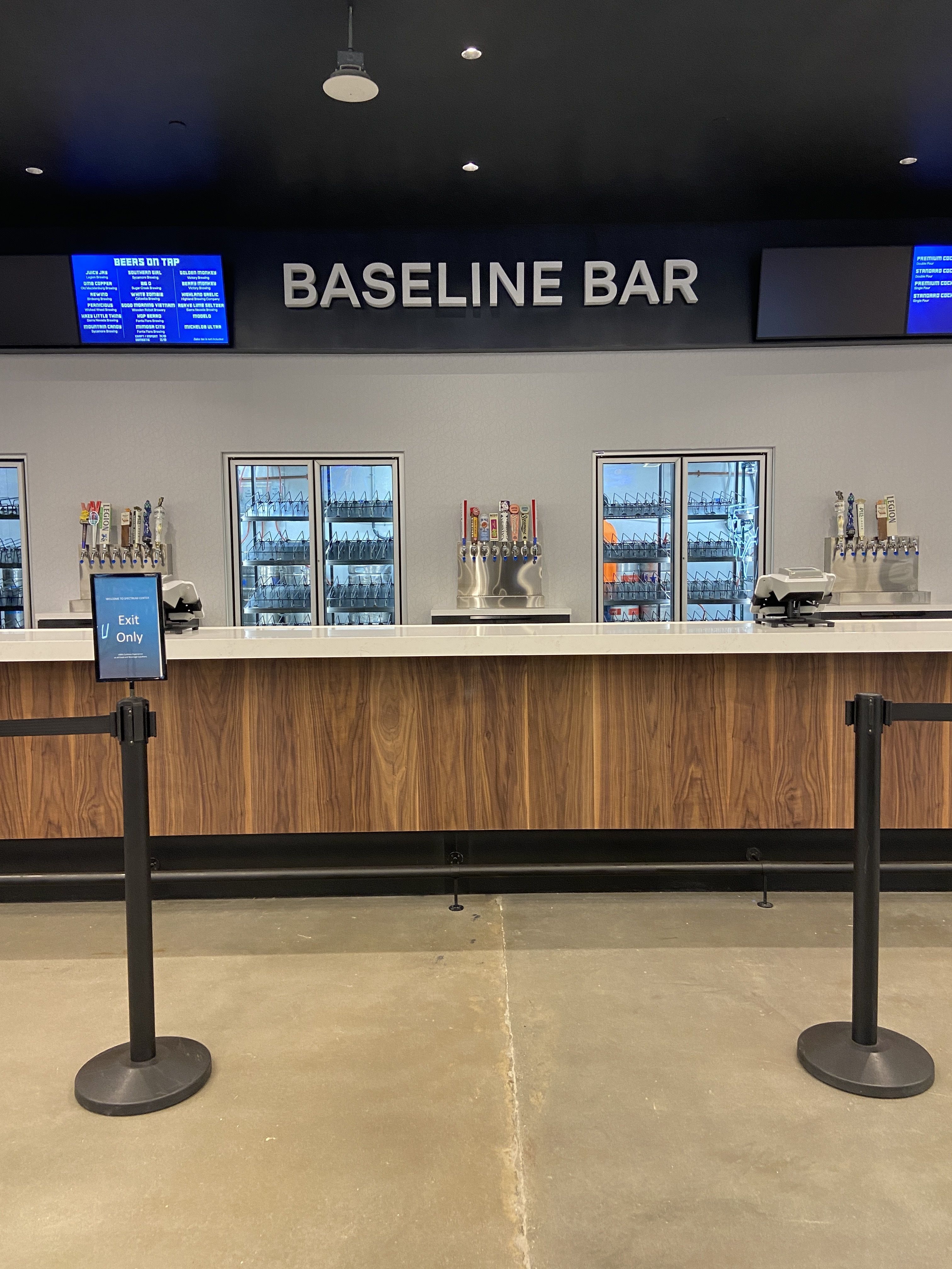 Empty bar counter with wooden panels and multiple beer taps under a sign reading "BASELINE BAR," with two digital menu screens displaying drink options.