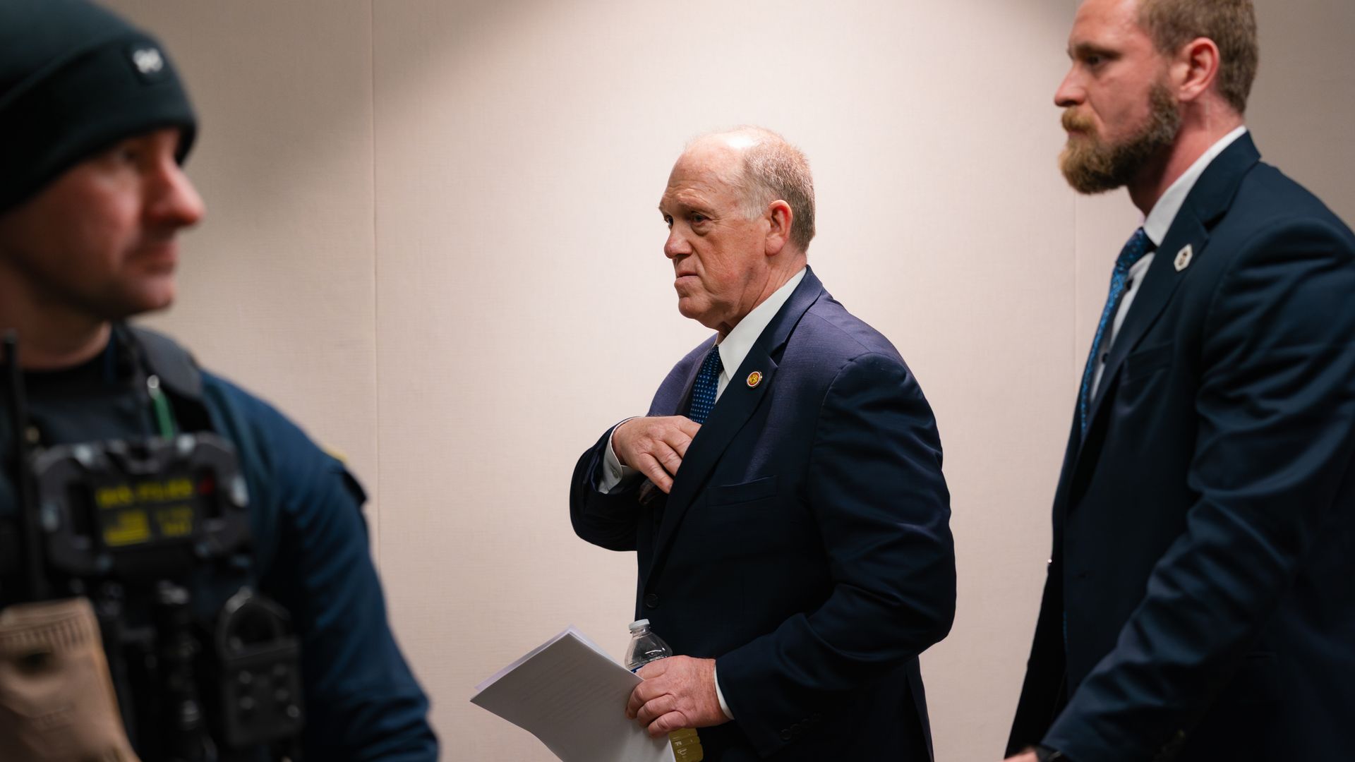Three men in suits and tactical gear walk indoors against a beige wall; middle man holds papers and a drink bottle, appearing serious and focused.