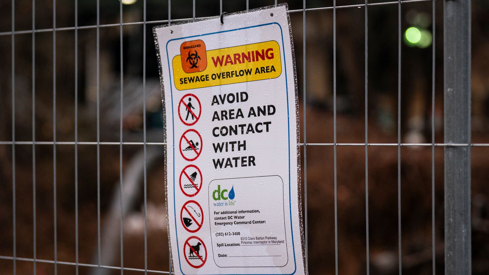 Warning sign on metal fence with biohazard symbol stating sewage overflow area. Advises to avoid area and contact with water. Includes DC Water emergency contact info.