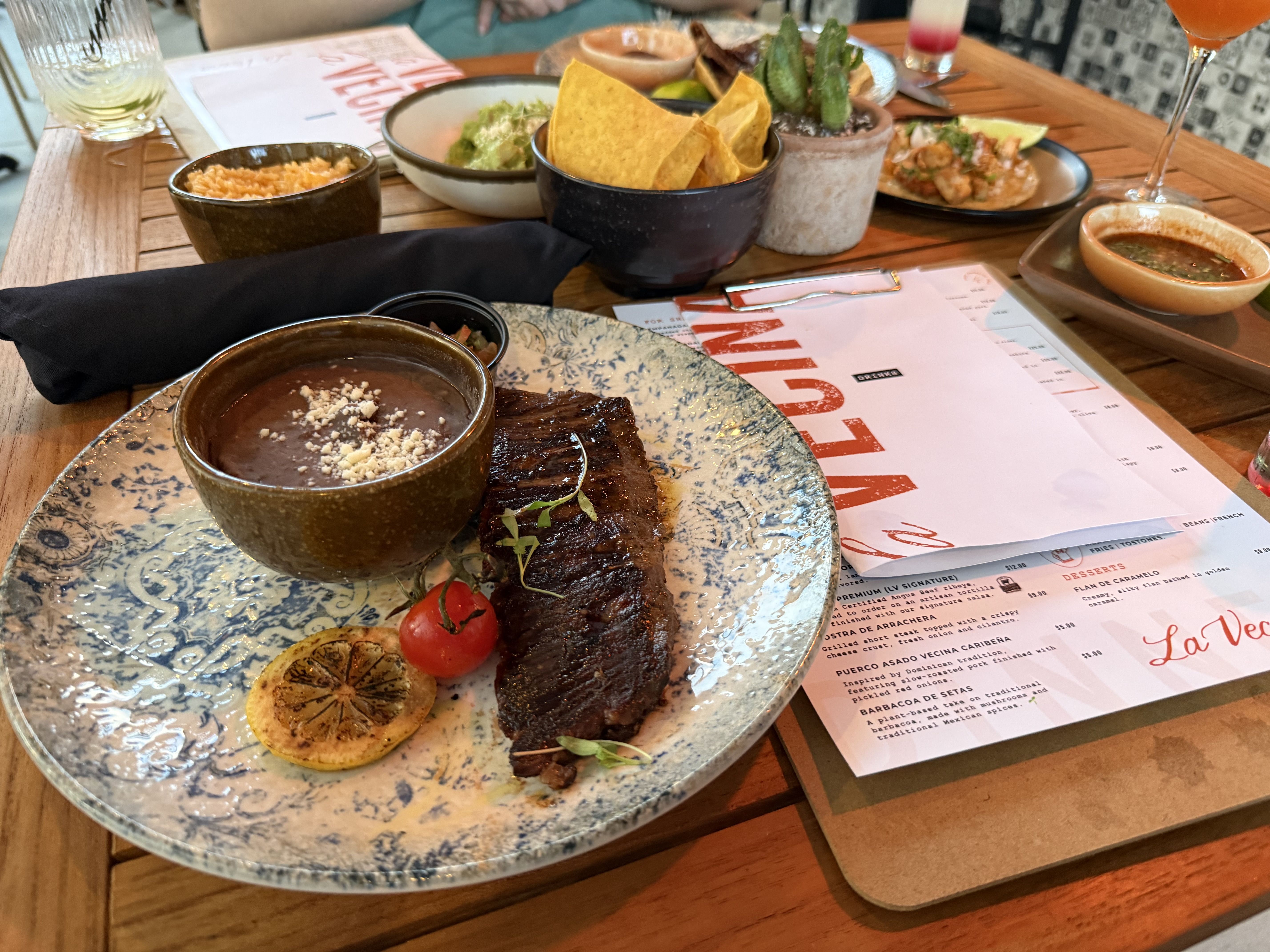 Wood table with a blue patterned plate holding a grilled steak with brown sauce, lemon slices, and a cherry tomato. A bowl of chips, a small sauce cup, guacamole, drinks, a plant, and a menu.