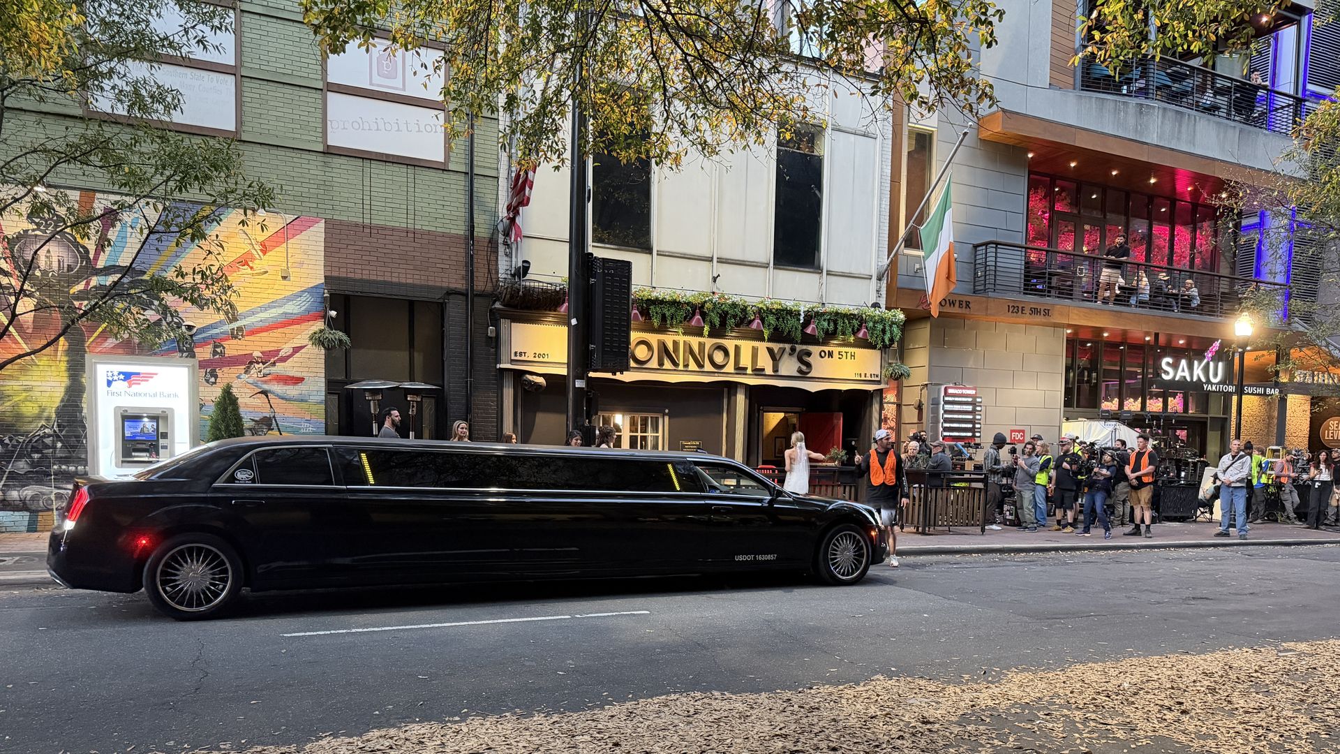 A long black limousine occupies a city street as a crowd lines the sidewalk outside Connolly's, with an Irish flag, a colorful mural on the wall, and autumn leaves scattered on the pavement.