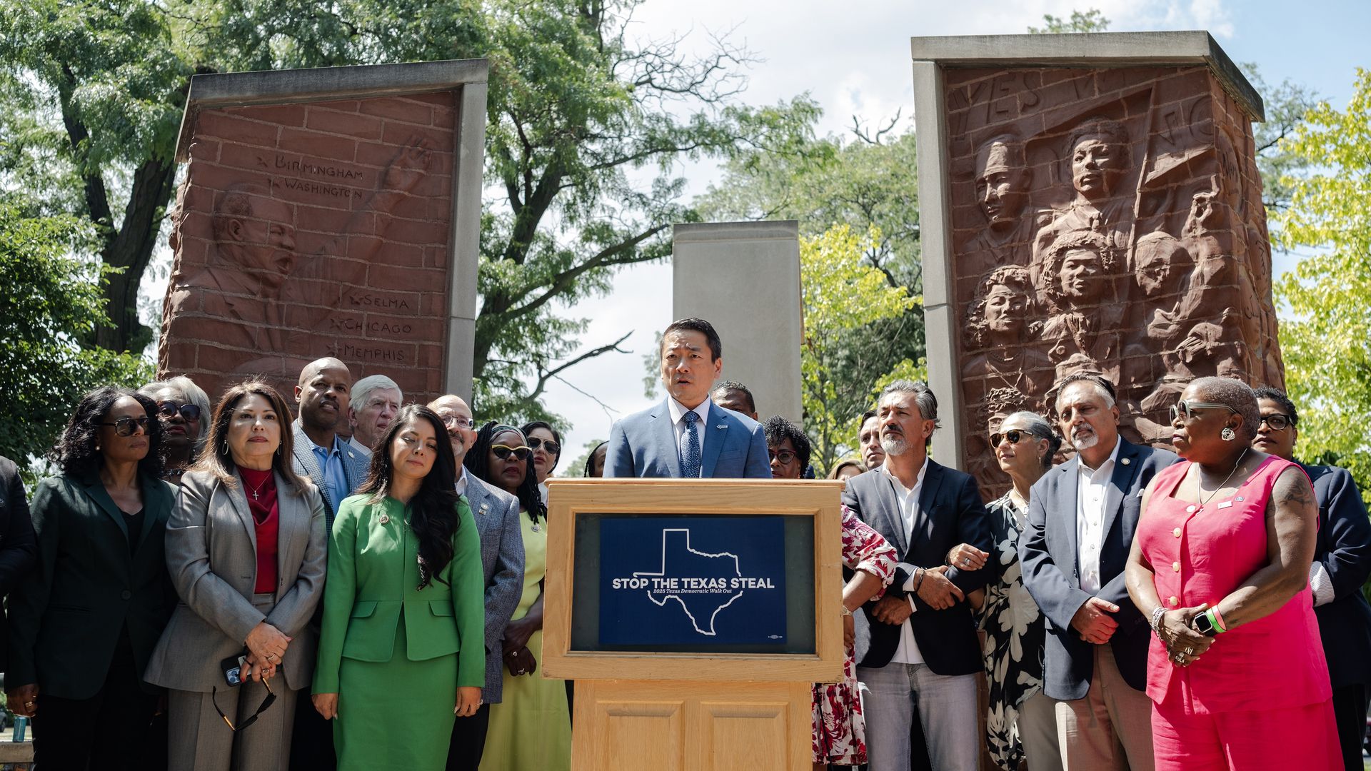 Diverse crowd outdoors in front of red brick relief columns; a man in a light blue suit speaks at a podium with a blue sign reading "Stop the Texas Steal".