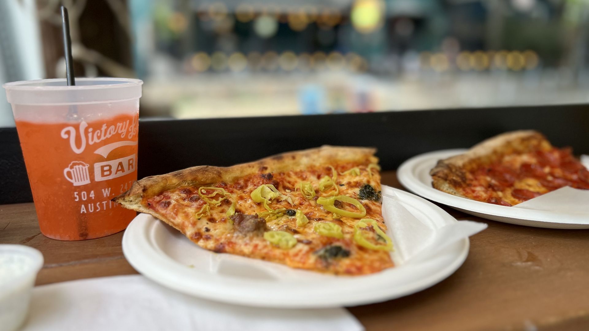 Two slices of pizza on white plates on a wooden table. One slice has banana peppers, the other pepperoni. An orange drink in a Victory Bar cup with a black straw is beside the pizzas.
