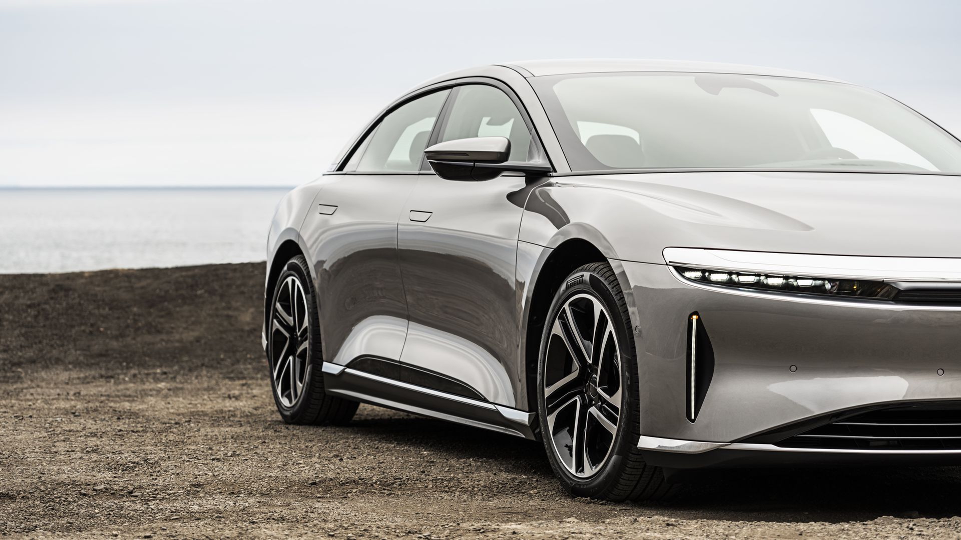 Gray electric car with sleek design and unique front LED lights parked on a dirt ground near a calm body of water under a cloudy sky.