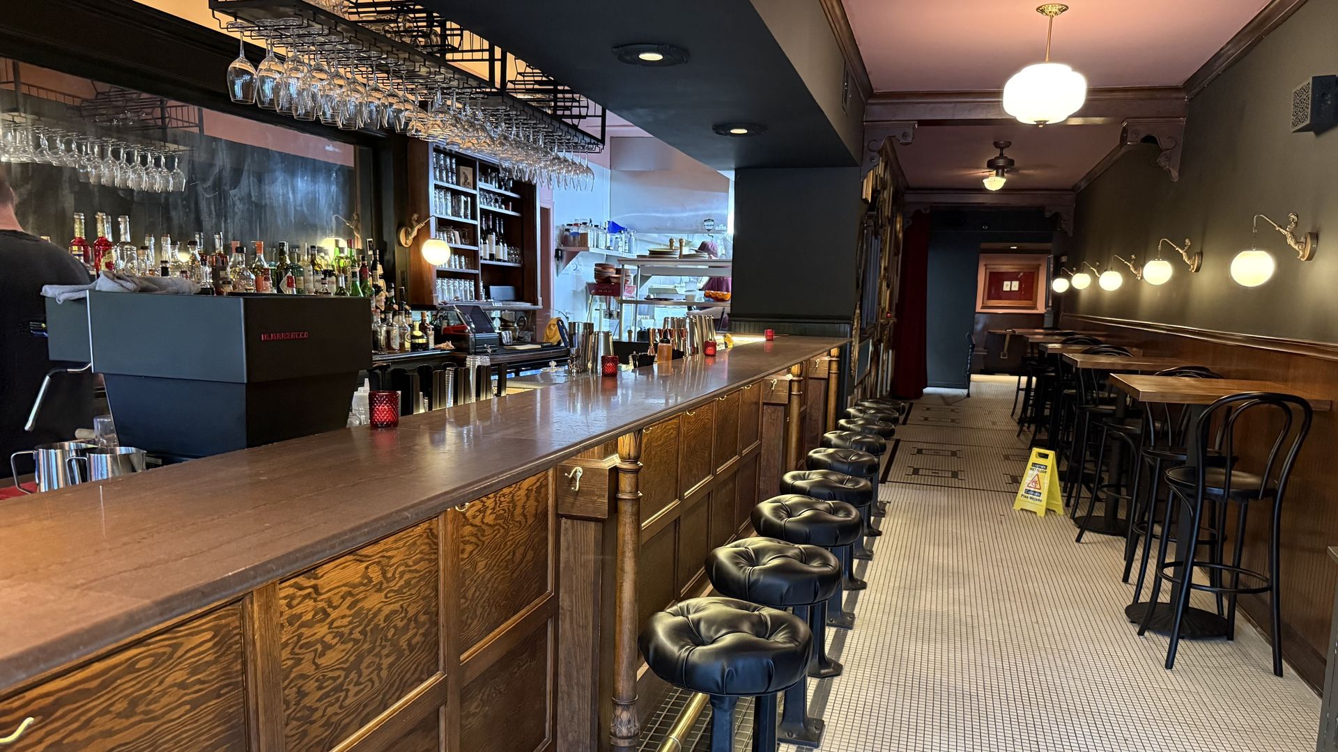 Long wooden bar with black stools, glass racks overhead, and liquor shelves. Warm lighting, open kitchen in back, tiled floor, and tables along the wall; caution sign on the floor.