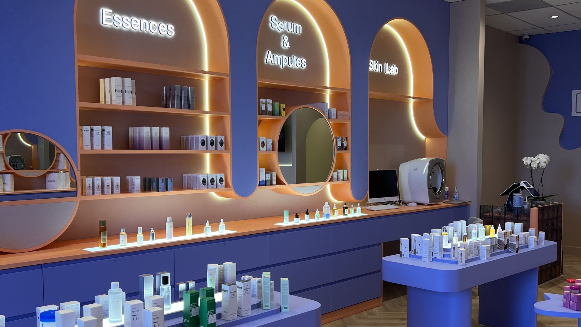 Interior of a skincare store with blue and orange shelves labeled "Essences," "Serum & Ampules," and "Skin Lab," wooden floor, and tables displaying various skincare products.