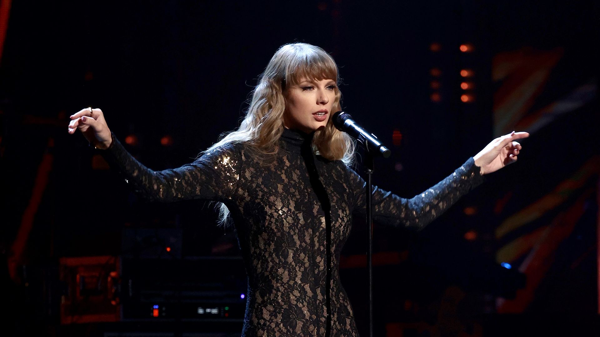 Taylor Swift in a black lace bodysuit performing on stage with arms extended, singing into a microphone against a dark, blurred background with red and orange lights.