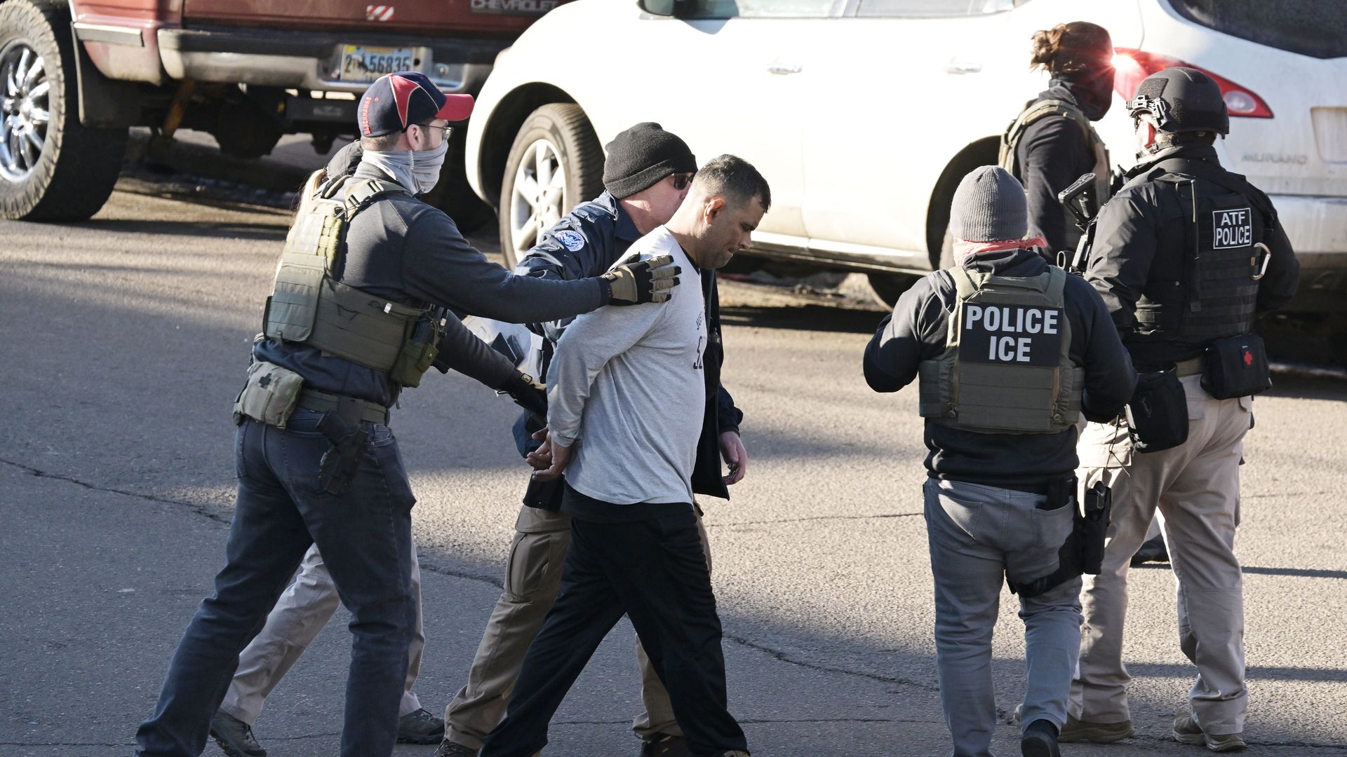 An immigrant in a white T-shirt is handcuffed and walked away by ICE agents