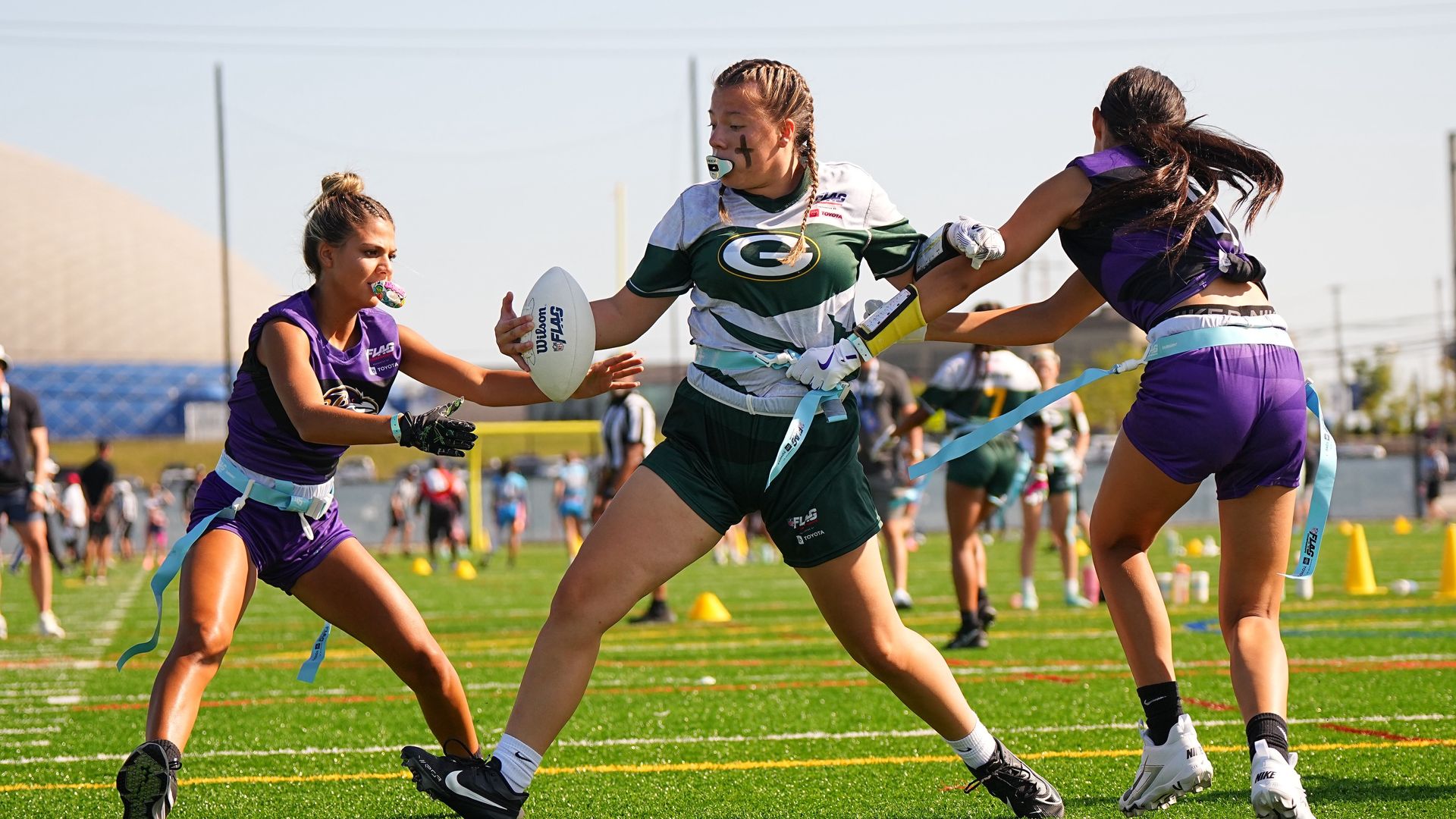 Three female flag football players on a sunny field. Center in green and white jersey with braided hair carries a Wilson football while two purple-uniform opponents reach to tackle.