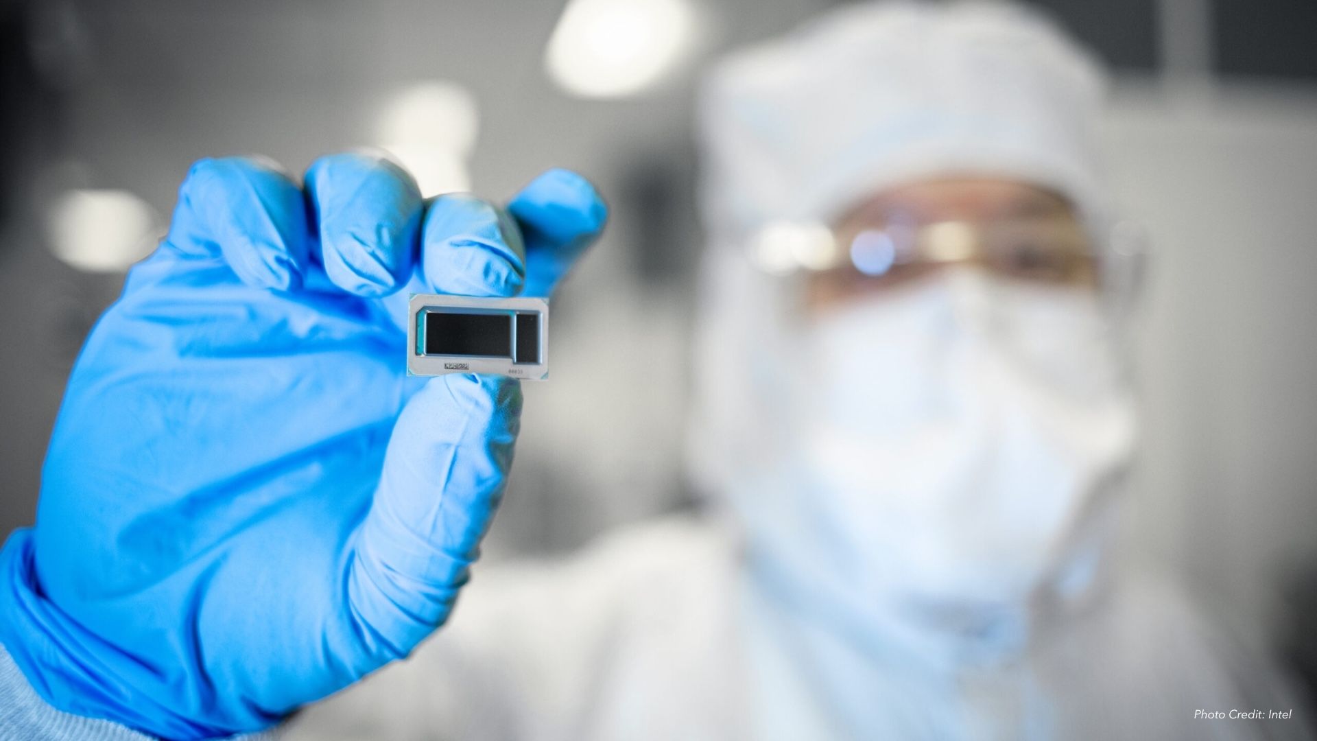 Scientist in white protective suit and blue gloves holding a small rectangular electronic chip in a lab setting with blurred background.