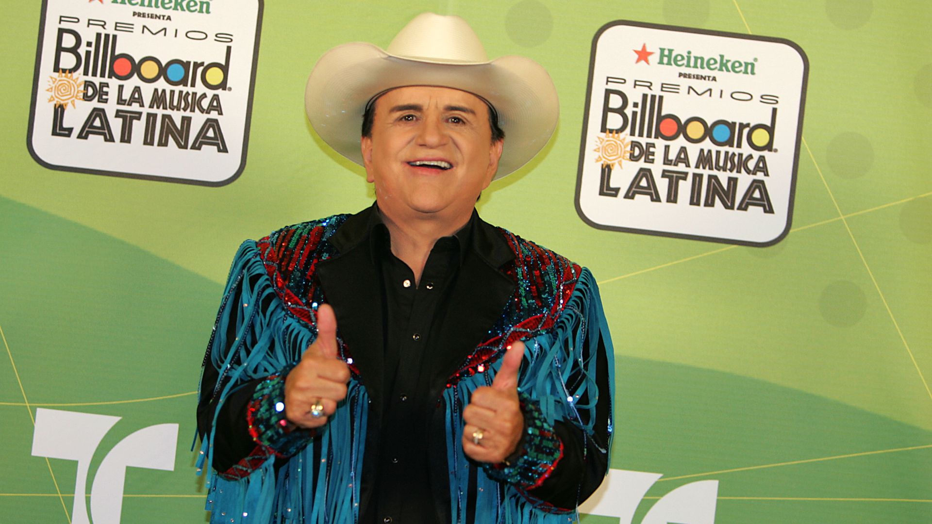 A man wearing a cowboy hat and fringed jacket smiles while holding his thumbs up in front of a green step-and-repeat.