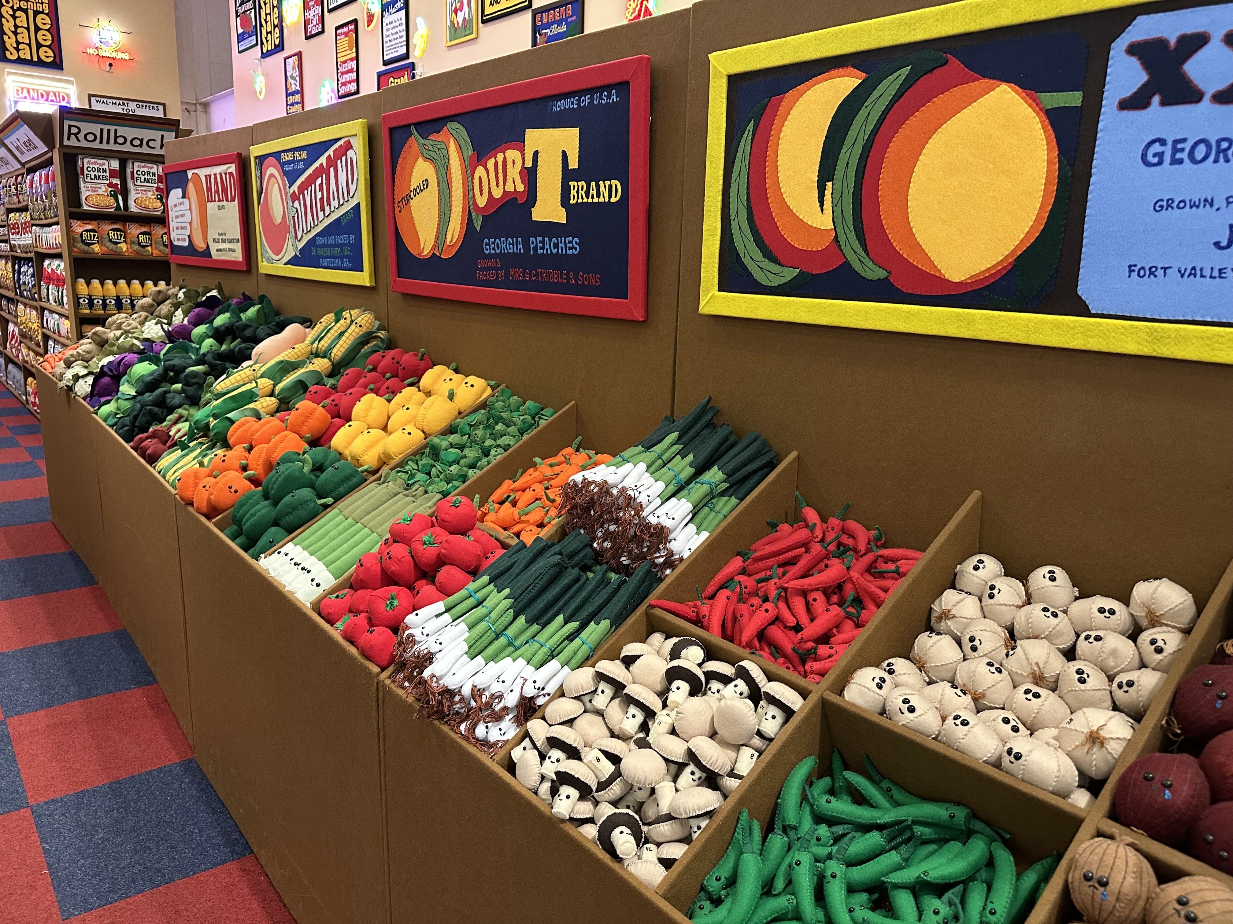 Colorful toy shop aisle with large felt fruit panels on the wall and long bins of plush produce (peaches, corn, peppers). Background shelves show cans and signs.