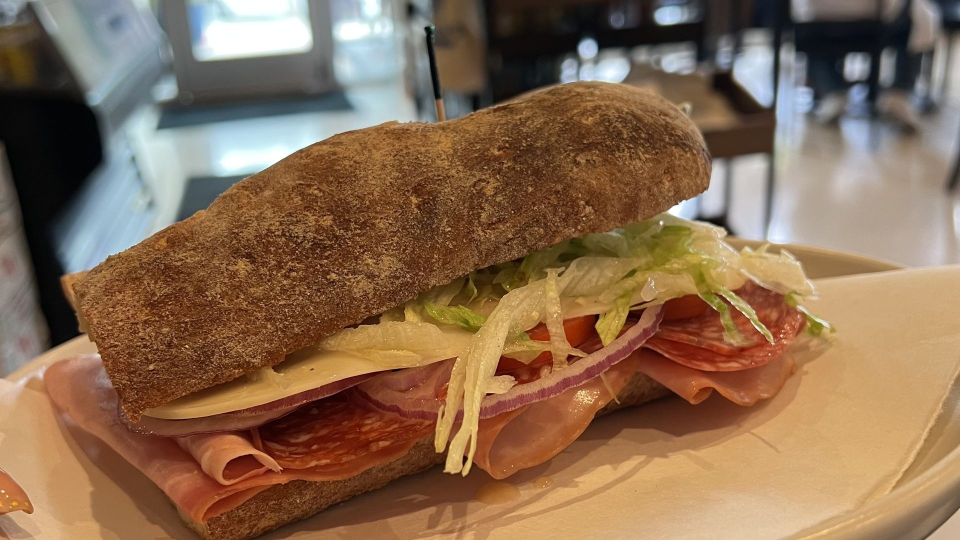 Close-up of a long, crusty brown bread sandwich filled with lettuce, tomato, and deli meats, served on a white plate in a casual cafe; a toothpick holds the sandwich together.