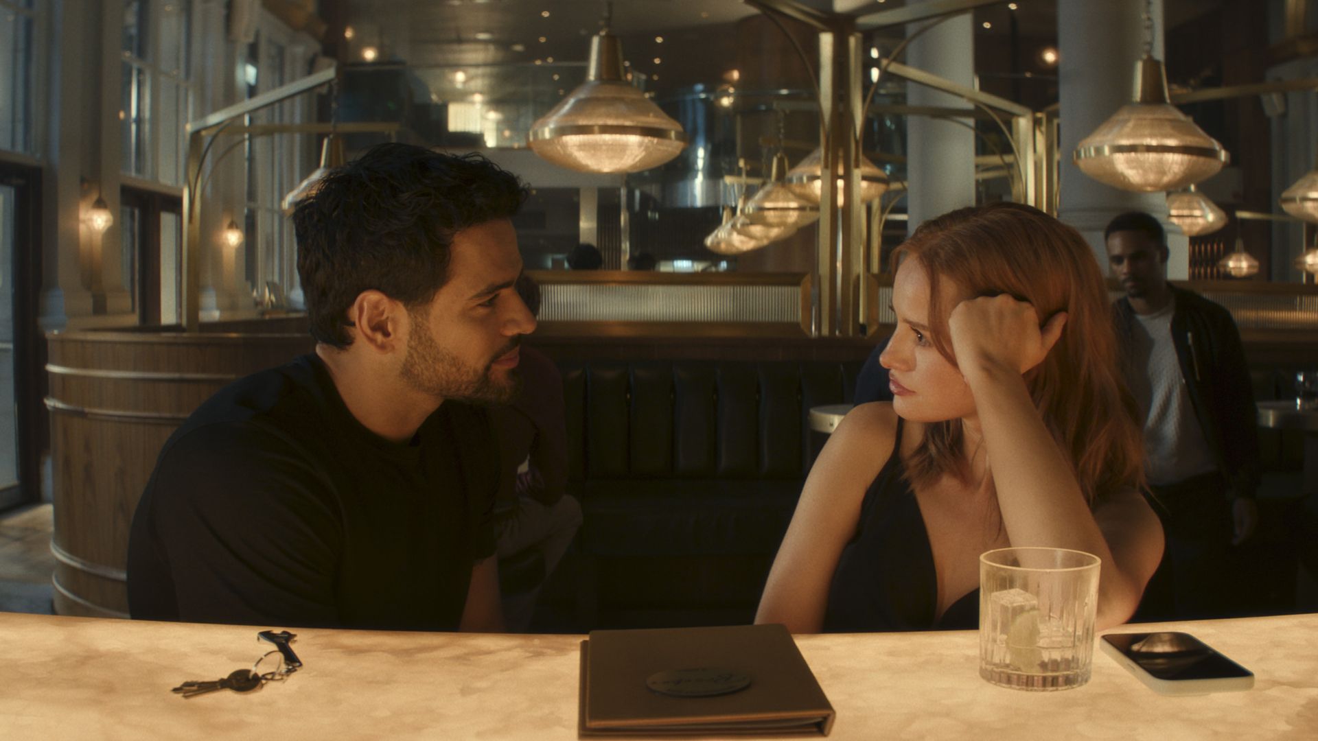 A man in a black shirt and a woman with red hair sit facing each other at a warmly lit bar with keys, a menu, a glass with ice, and a phone on the counter, inside a stylish restaurant.
