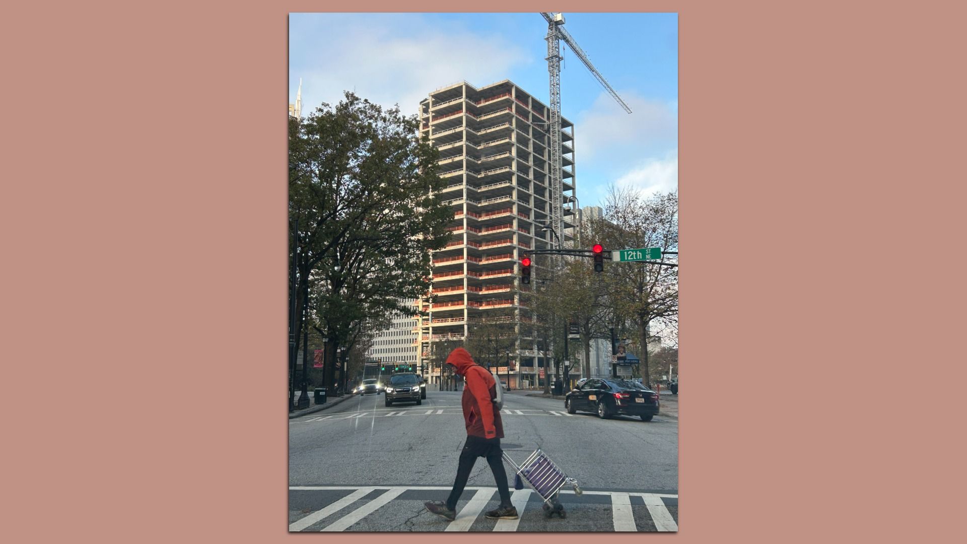 Unfinished concrete building with a tower crane rises over a city street. A man in an orange hoodie crosses the crosswalk pulling a striped suitcase; red light and trees along the road.