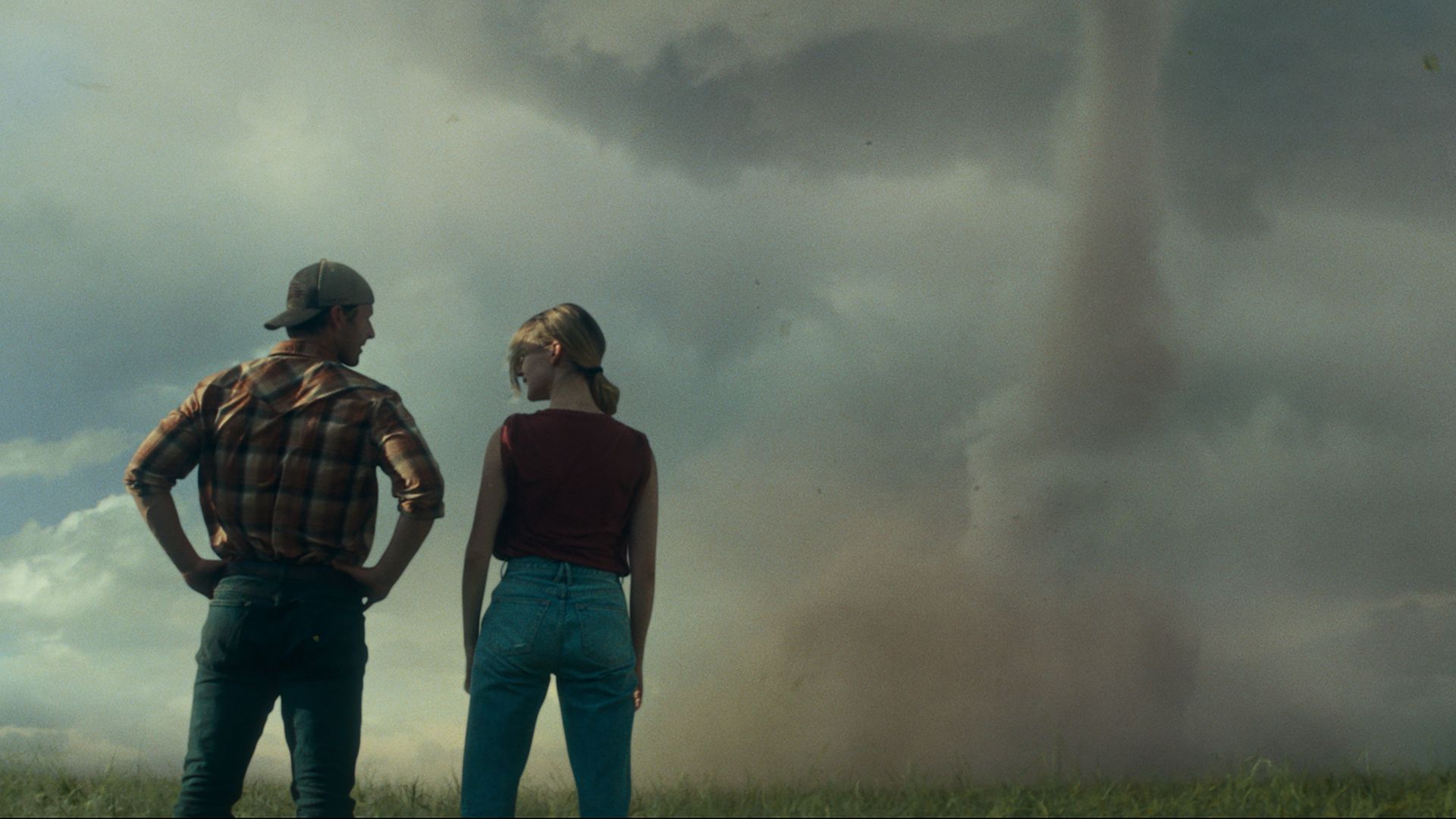 Two people stand in front of a stormy sky with a tornado. 