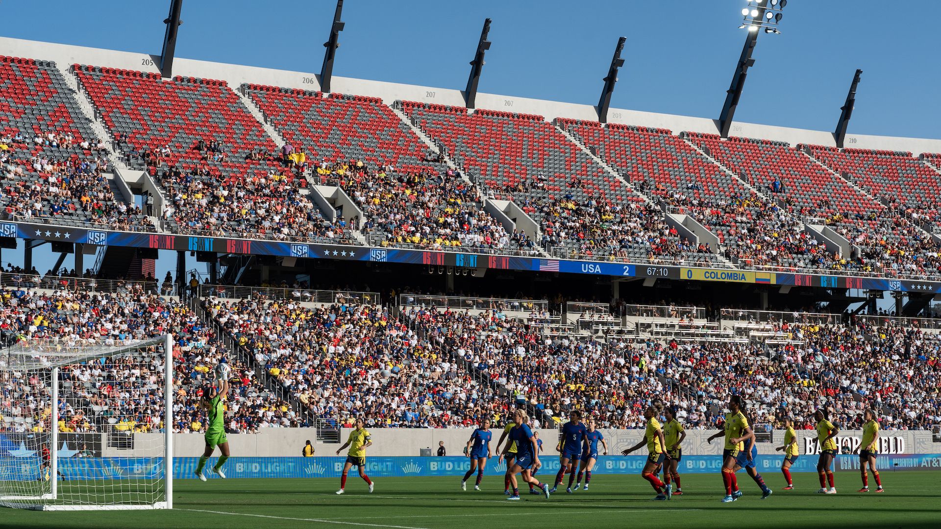 The U.S. women's national team players in blue jerseys attack the goal during a game vs. Colombia in yellow jerseys at Snapdragon Stadium with fans in the stands.