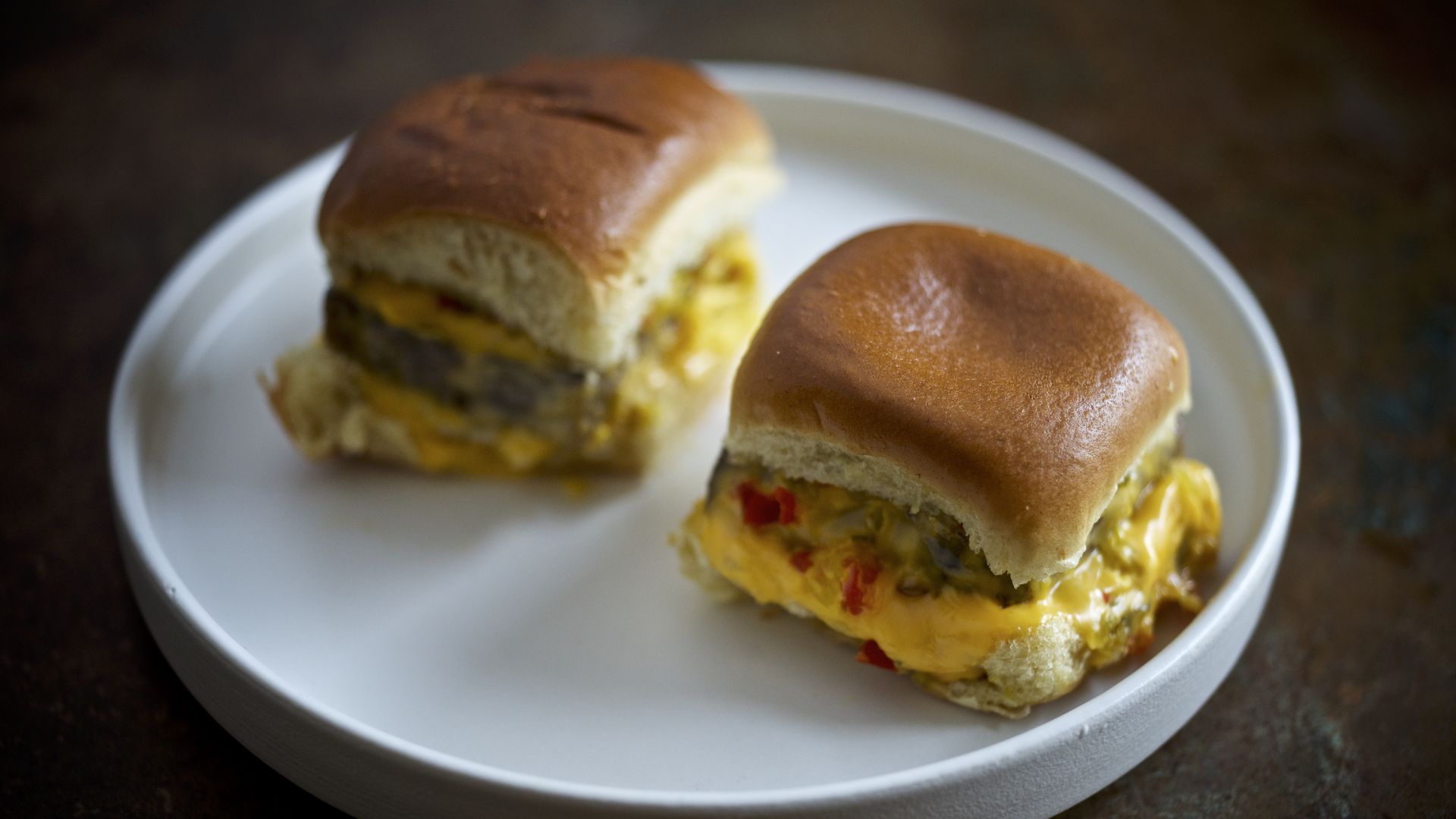 Two cheeseburger sliders with melted yellow cheese and bits of red pepper on soft square buns, served on a white round plate against a dark background.