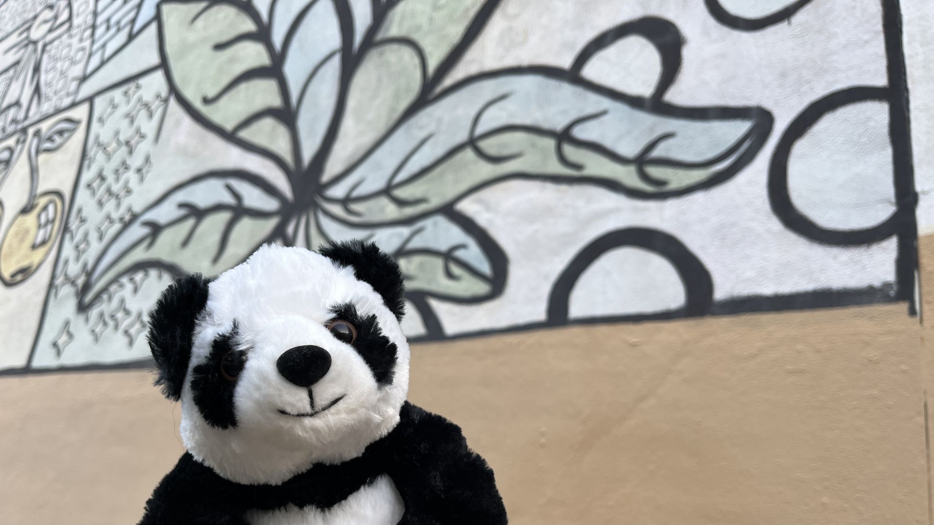 A stuffed animal panda is held in front of a faded mural with leaves and cherries.
