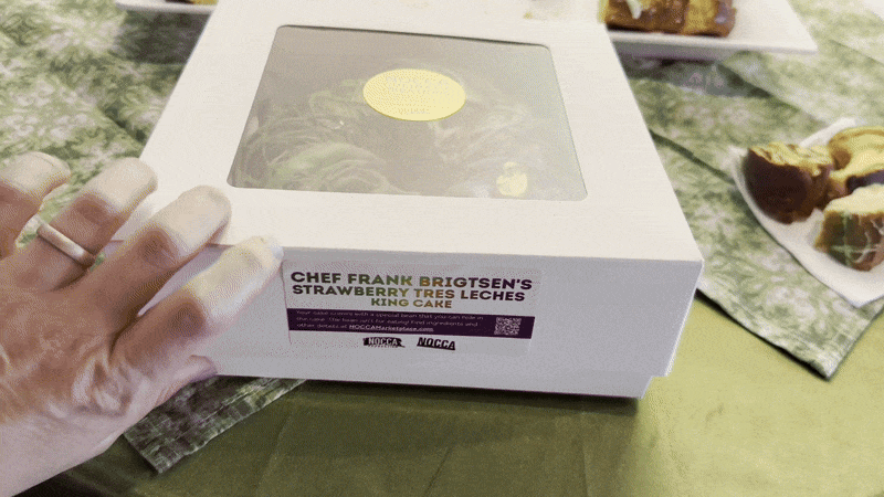 A hand reaches to open a cake box with a purple, green and gold-decorated king cake inside. 