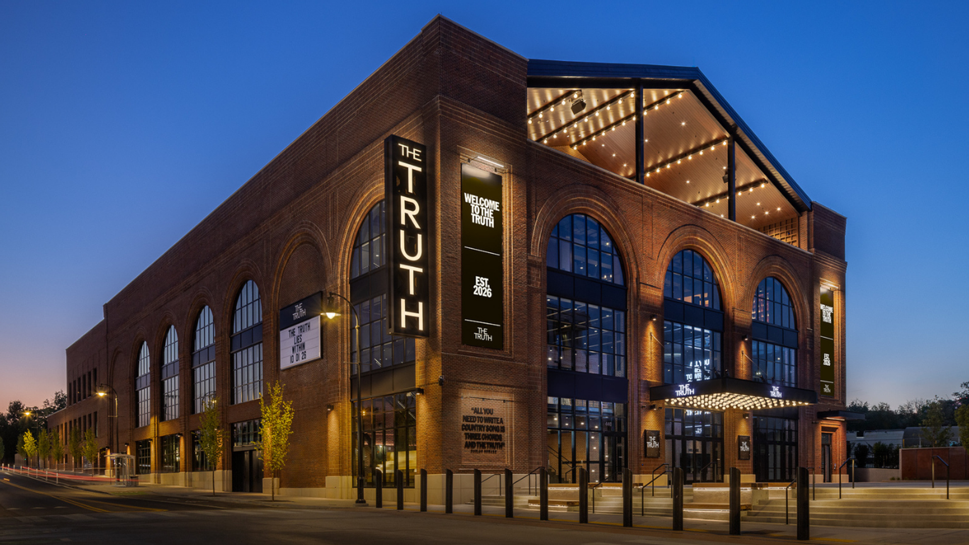 Brick theater at dusk with a tall vertical sign reading "THE TRUTH" beside arched windows. Upper glass level lit by string lights; banners read "WELCOME TO THE TRUTH."