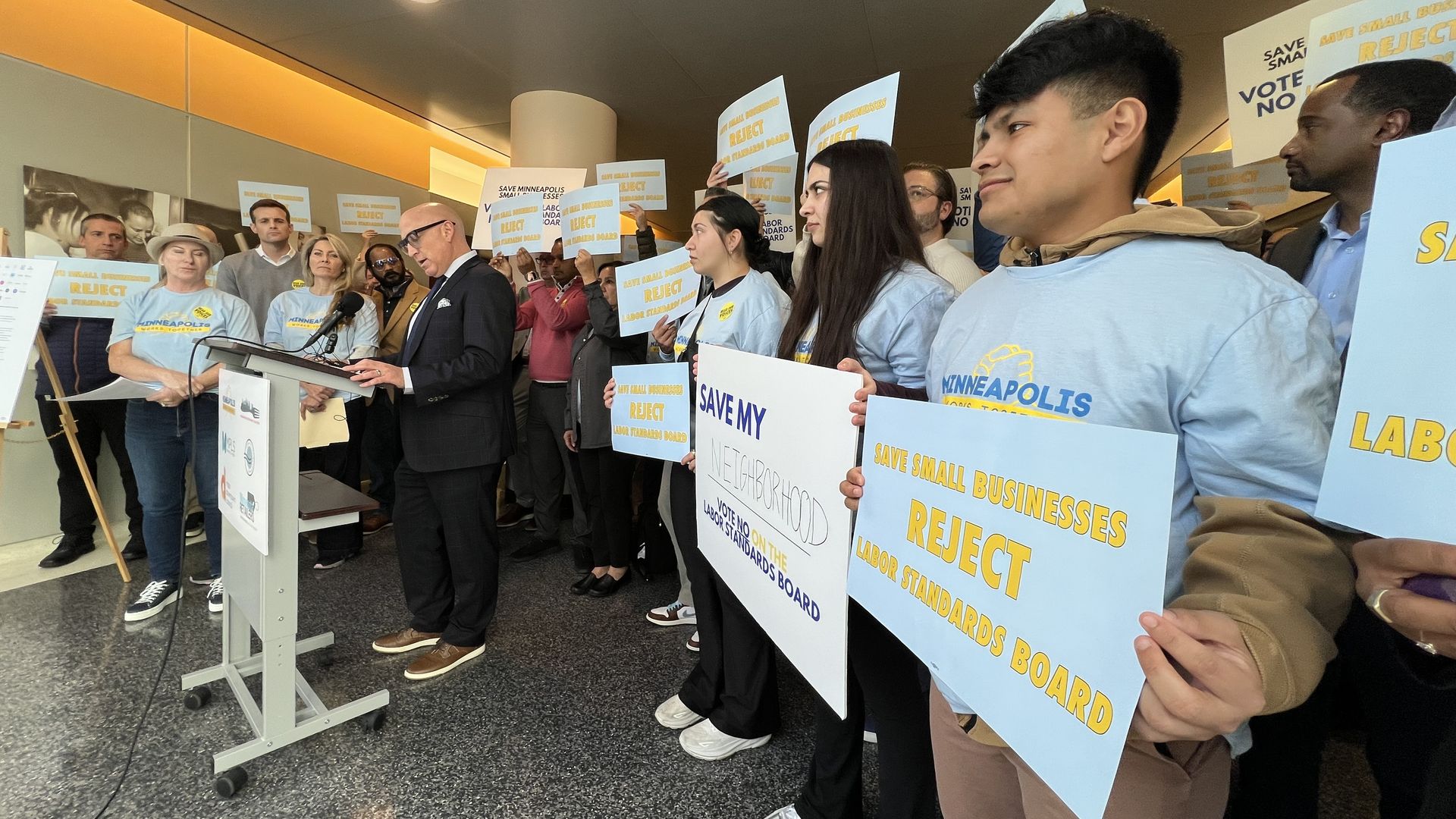 A crowd of people in light-blue shirts holds signs reading "Save Local Businesses, Reject Labor Standards Board" at a press conference