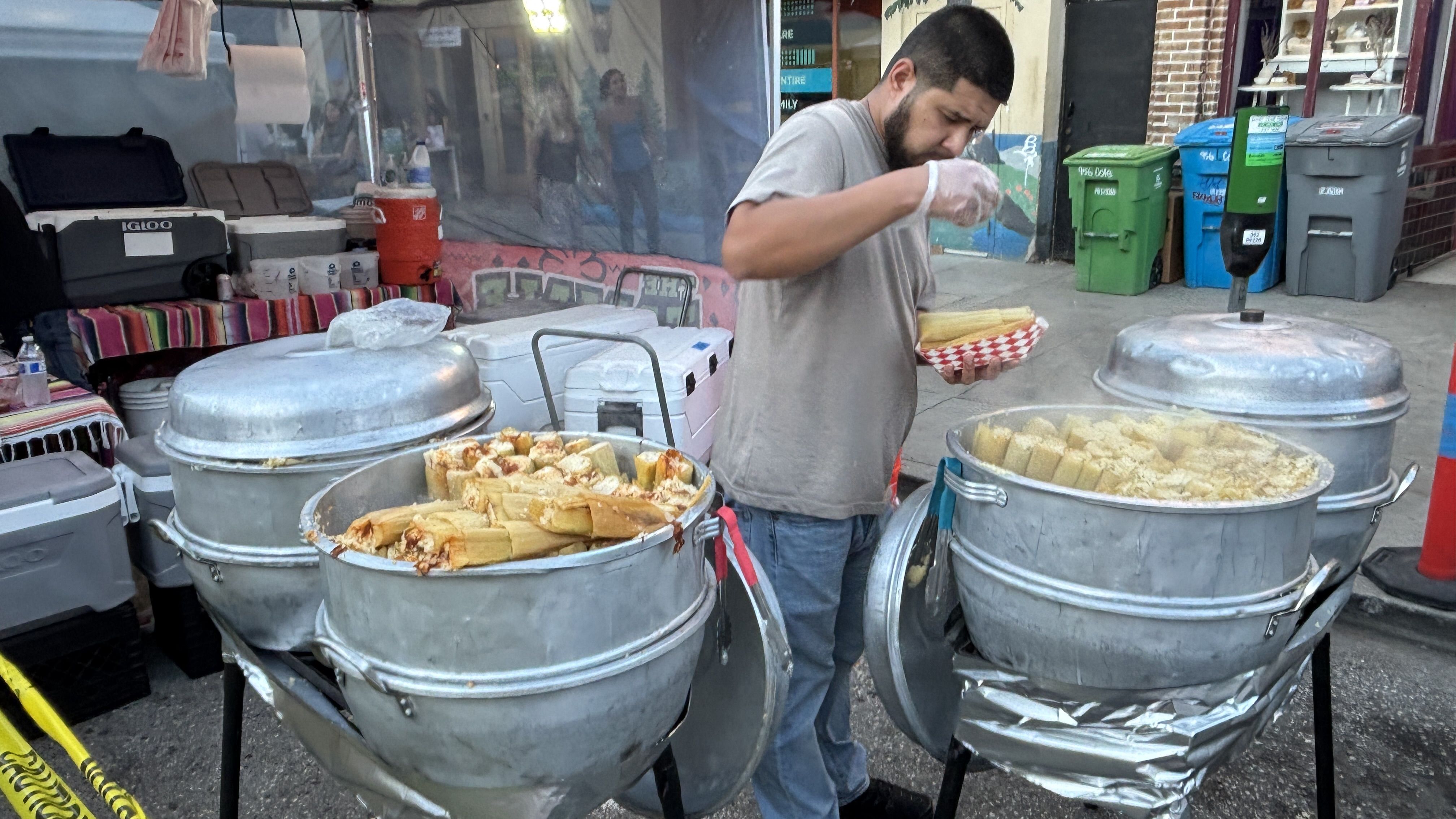 Street vendor in a gray T-shirt and gloves scoops food from three large metal steamers into a red-and-white checkered basket, with coolers and city bins in the background.