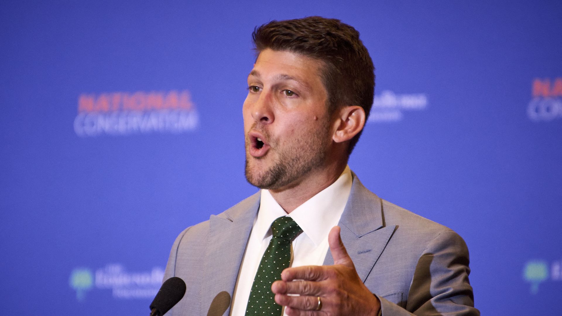 A man with brown hair in a light blue suit, green-and-white polka dot die and white button-down shirt motions with his right hand while speaking into a microphone. A blue background behind him reads "NATIONAL CONSERVATISM."