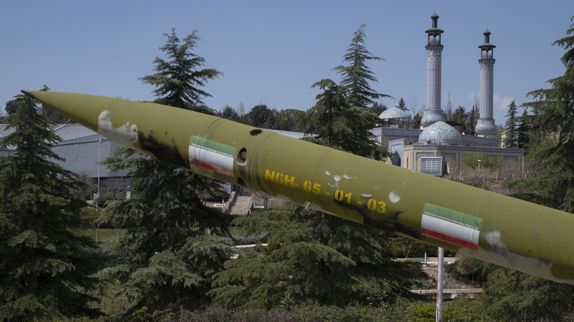 An outdoor museum scene with a large green missile angled across the foreground and ornate blue-tiled minarets and a domed building in the background, amid trees under a blue sky.