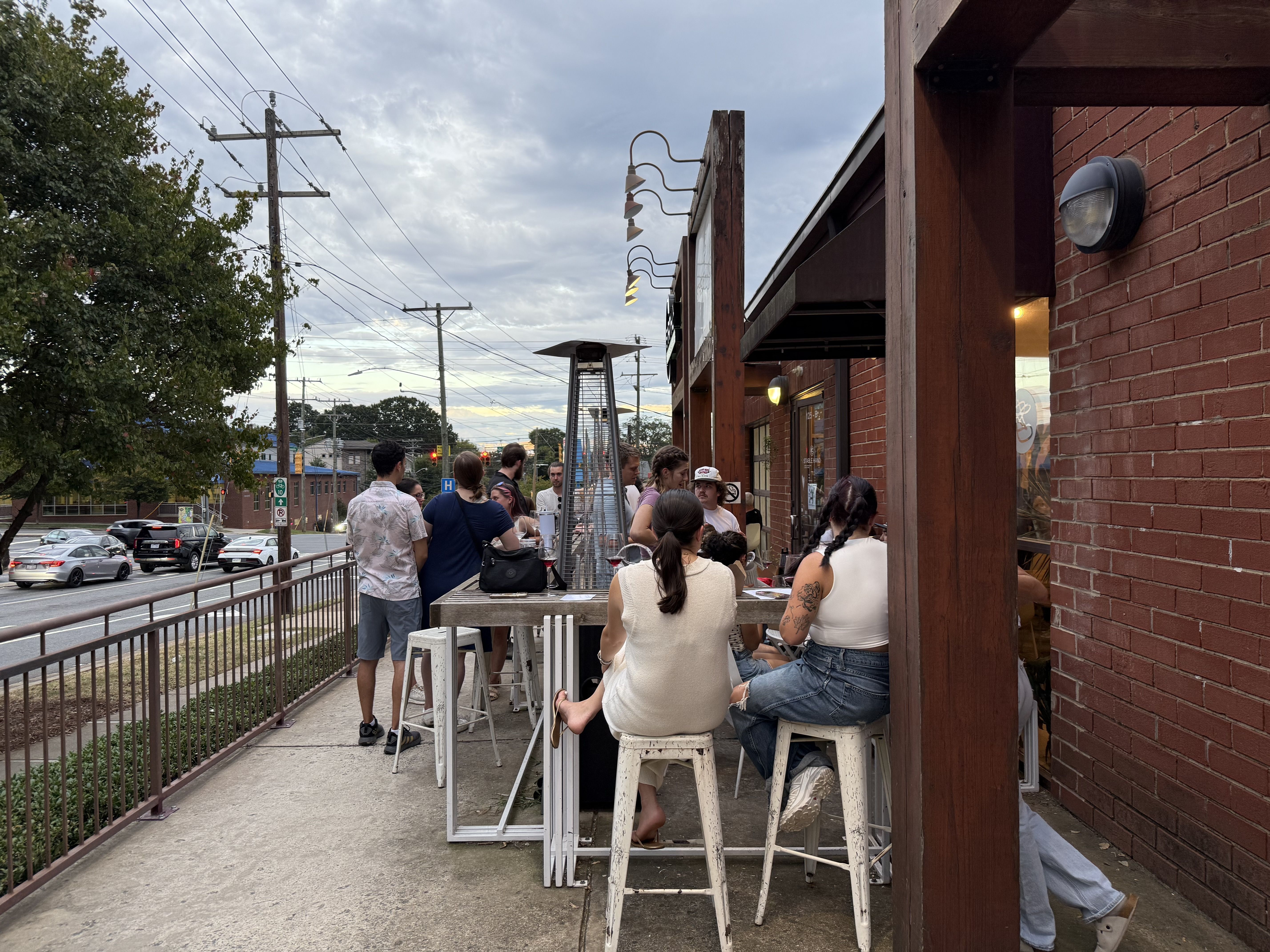 People socializing at outdoor bar seating with tall stools, heater, and brick building under cloudy sky near a street with cars and power lines.