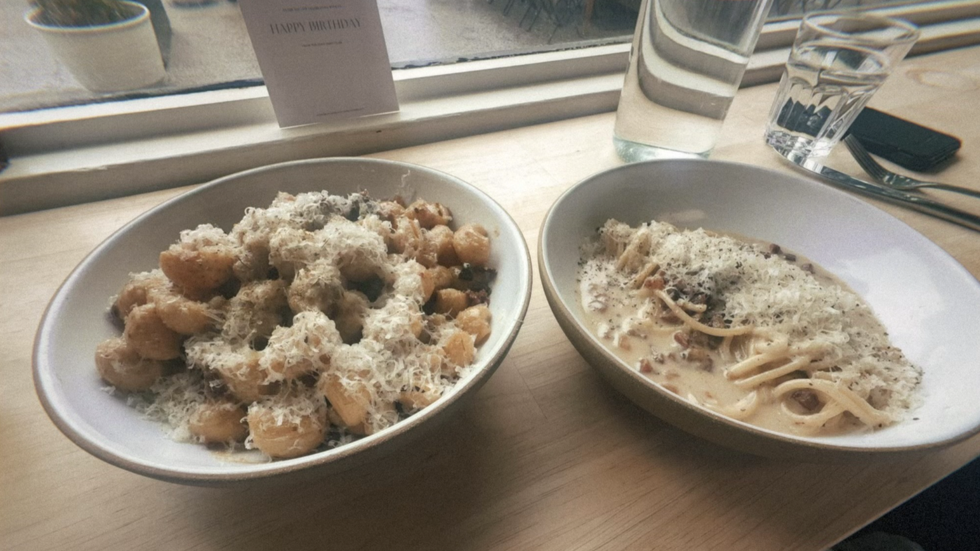 Two bowls of pasta on a light wooden table by a window: left gnocchi with grated cheese, right creamy fettuccine with mushrooms, topped with cheese; a "Happy Birthday" card in the background.