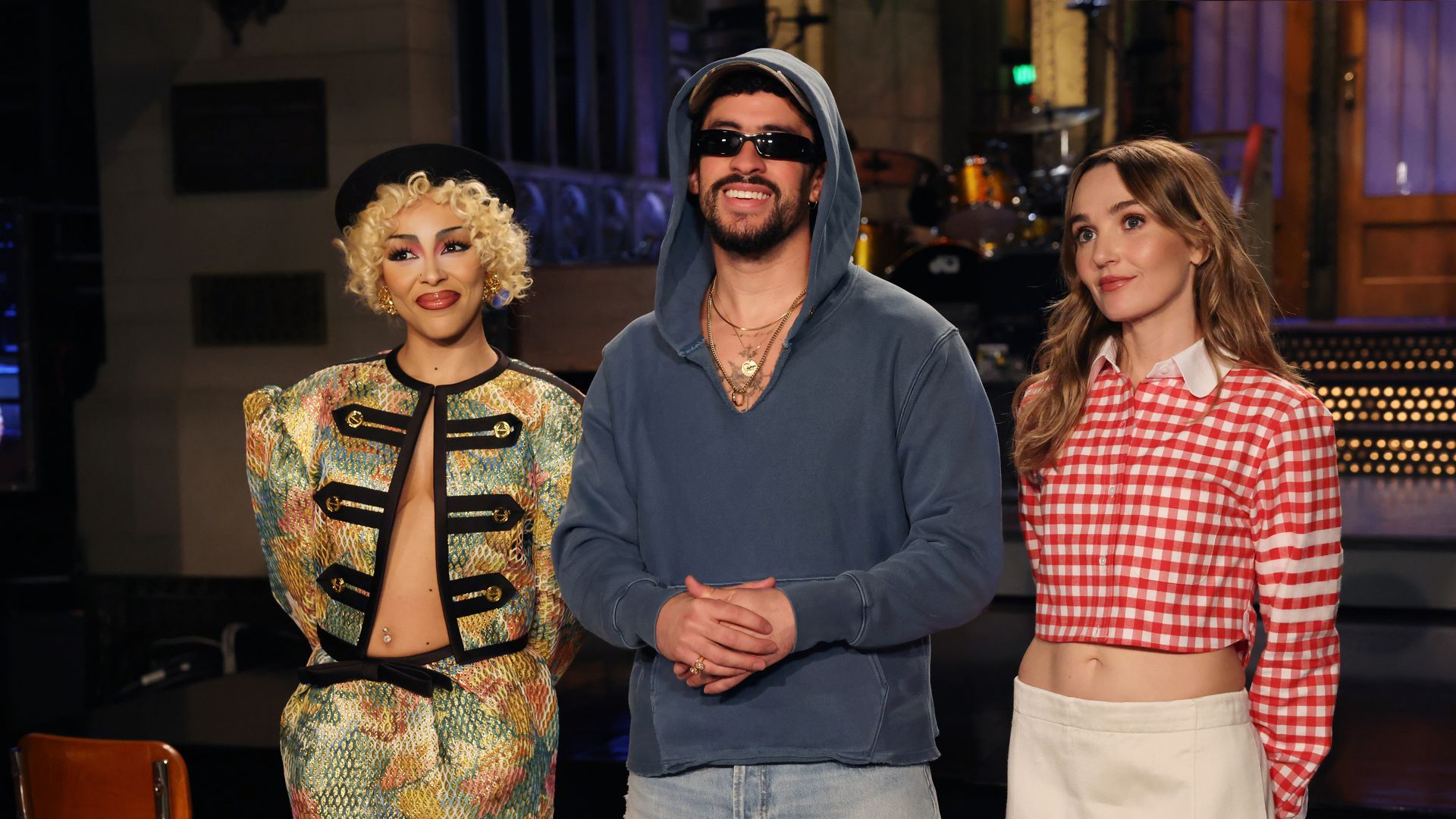Doja Cat, host Bad Bunny standi indoors with musical instruments in the background. Chloe Fineman Doja Cat on the left wears a colorful patterned outfit and black beret. Bad Bunny  wears a blue hoodie and sunglasses.  Chloe Fineman wears a red and white checkered cropped shirt and white skirt.