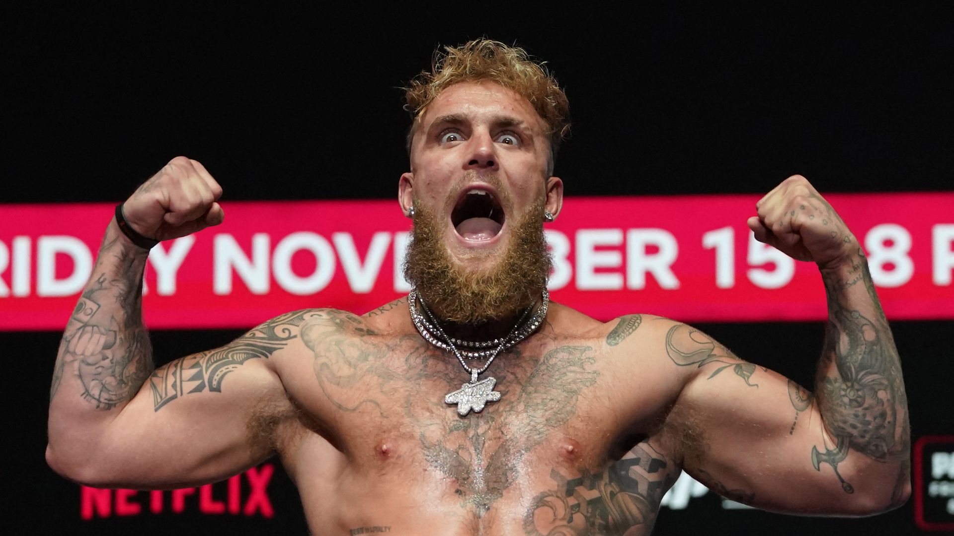 Shirtless man with tattoos and beard flexing his muscles and yelling, wearing multiple necklaces, with a red and black background showing partial date text and Netflix logo.