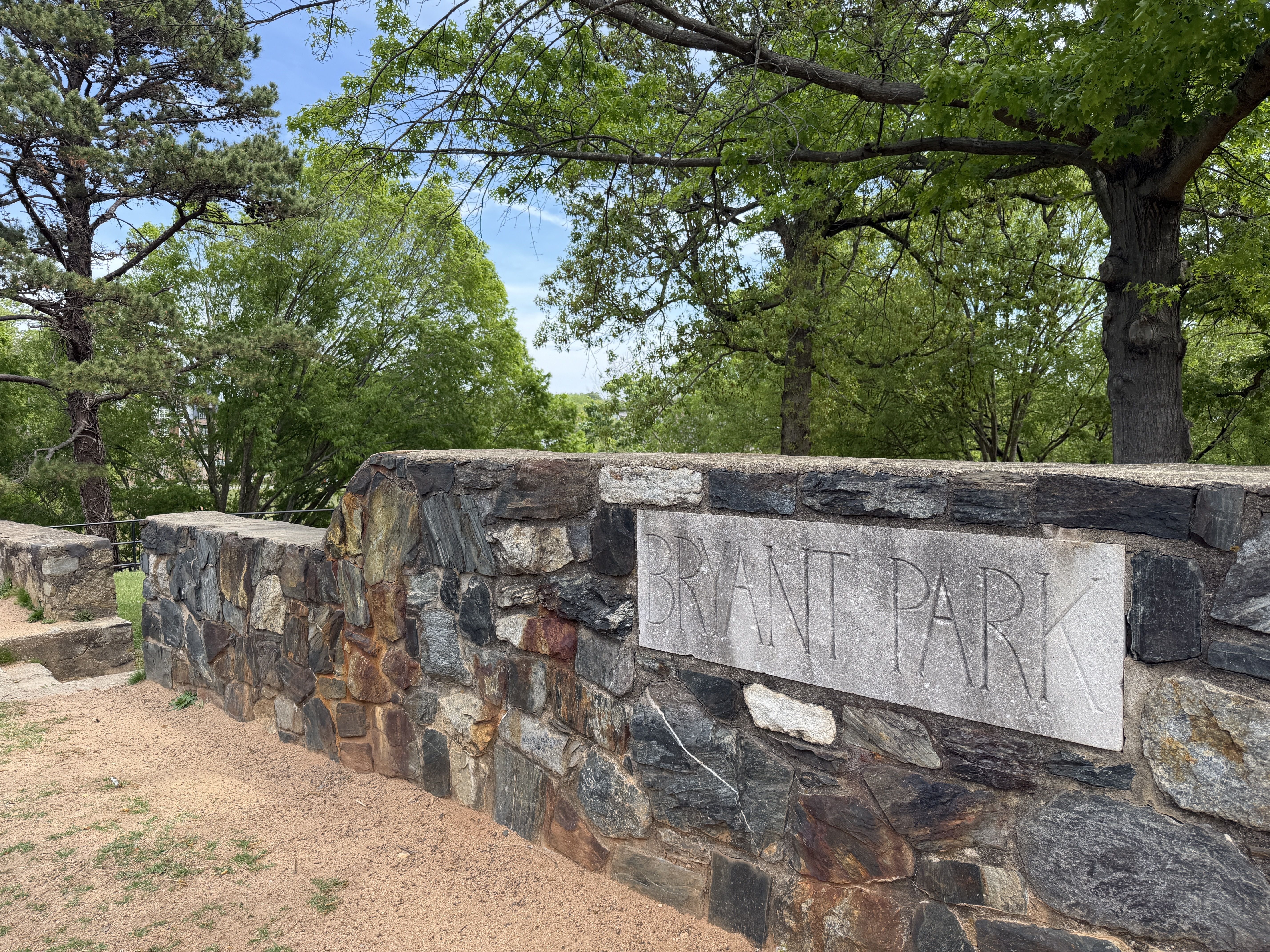 A stone wall with a rectangular gray plaque reading "BRANT PARK" sits beside a dirt path, surrounded by green trees under a bright blue sky.