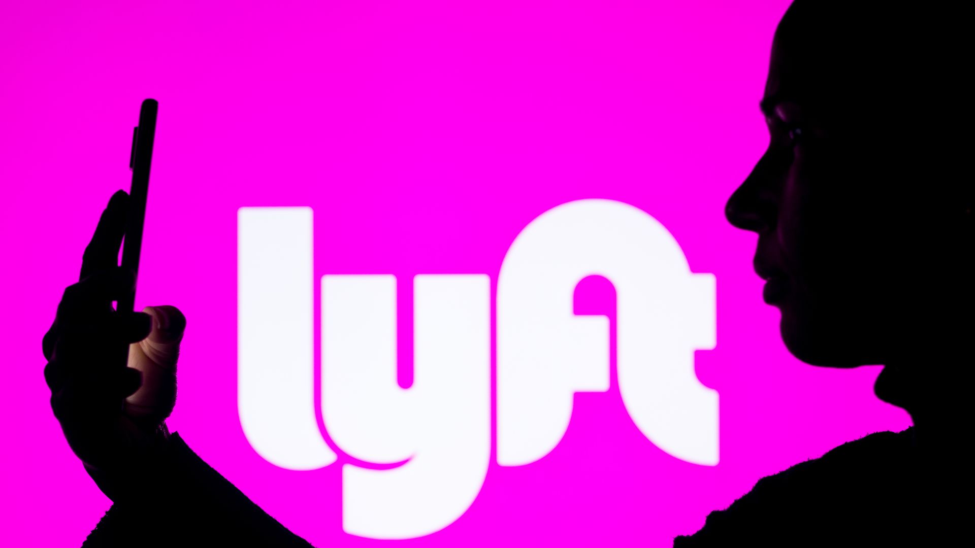 A photo illustration shows a silhouette of a woman in front of a neon pink Lyft sign, looking at her phone presumably to request a lyft