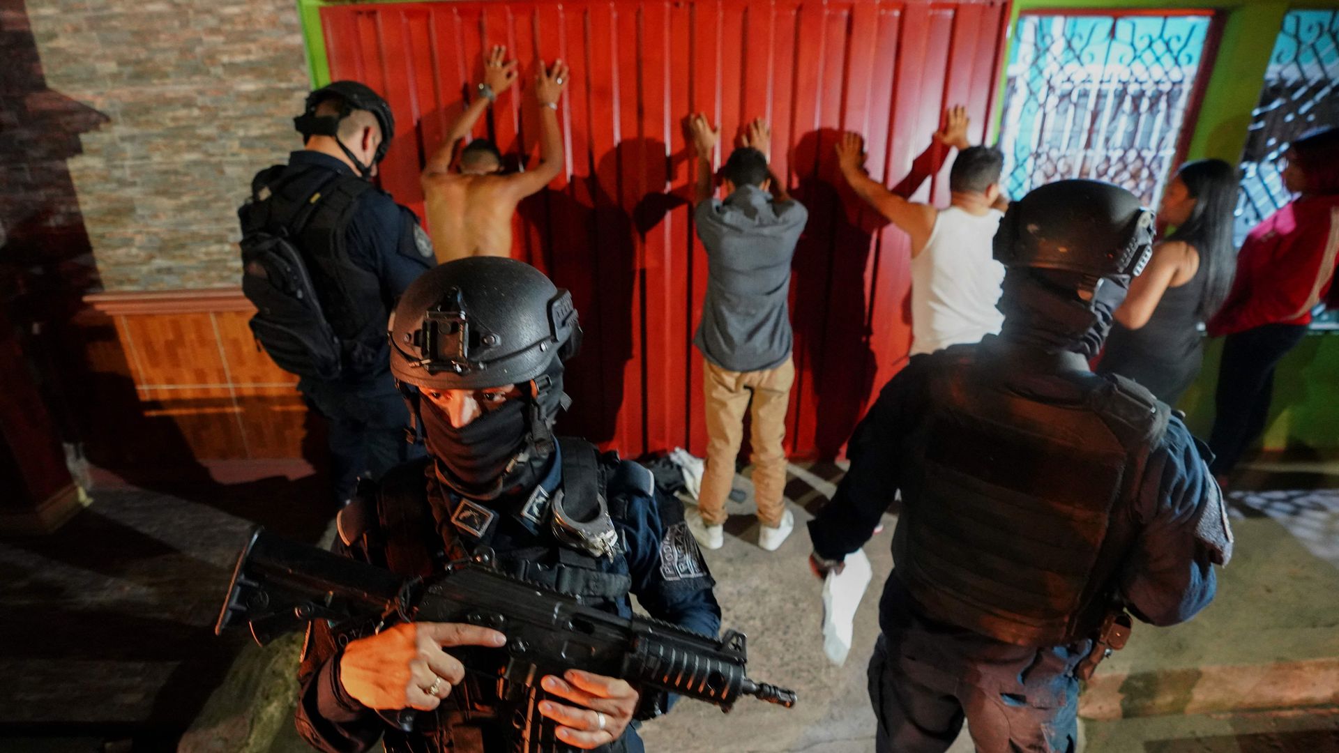 two soldiers in full tactical gear stand in front of a row of men who have their hands up against a red wall 