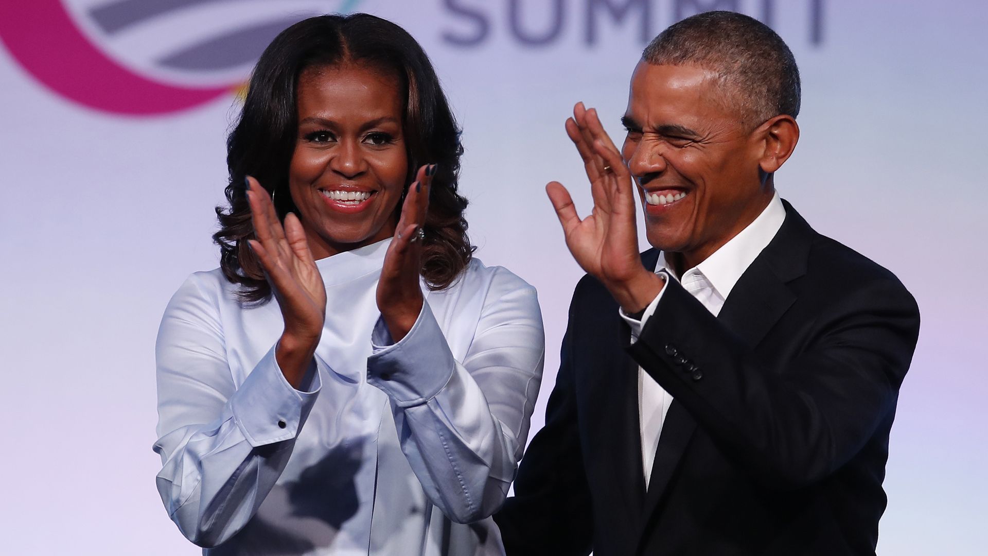 Barack and Michelle Obama smiling, with Michelle clapping and Barack waving.