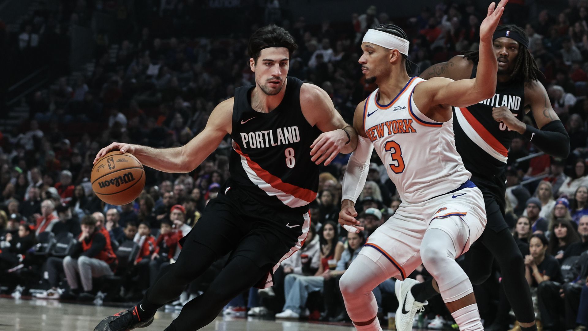 Portland Trail Blazers player in black jersey dribbling basketball while guarded by New York Knicks player in white jersey during a game, with another Portland player in background and crowd watching.