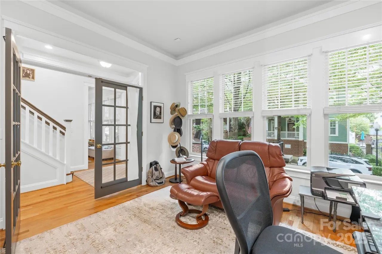 Bright home office with large windows, brown leather armchair, black mesh office chair, hardwood floors, and gray French doors leading to a stairway. Hats and bags hang on the wall.