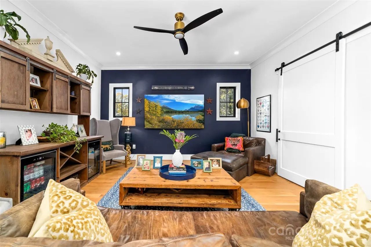 Cozy living room with tan leather couch, wooden coffee table with framed photos and flower vase, navy accent wall with landscape TV, two windows, gray wingback chair, and white sliding barn door.