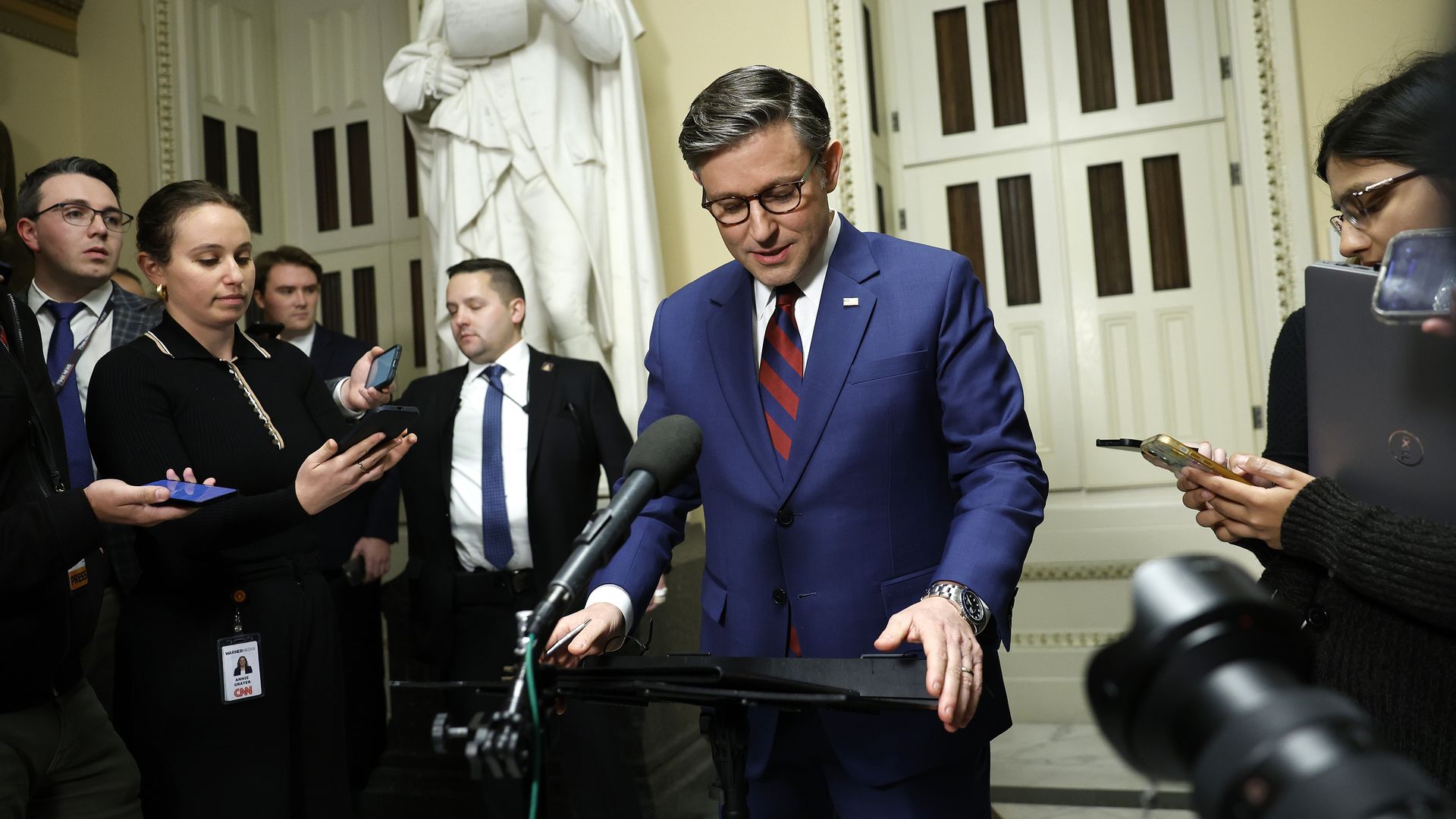 House Speaker Mike Johnson, wearing a blue suit and gripping a podium in front of a white wall and a marble statue.