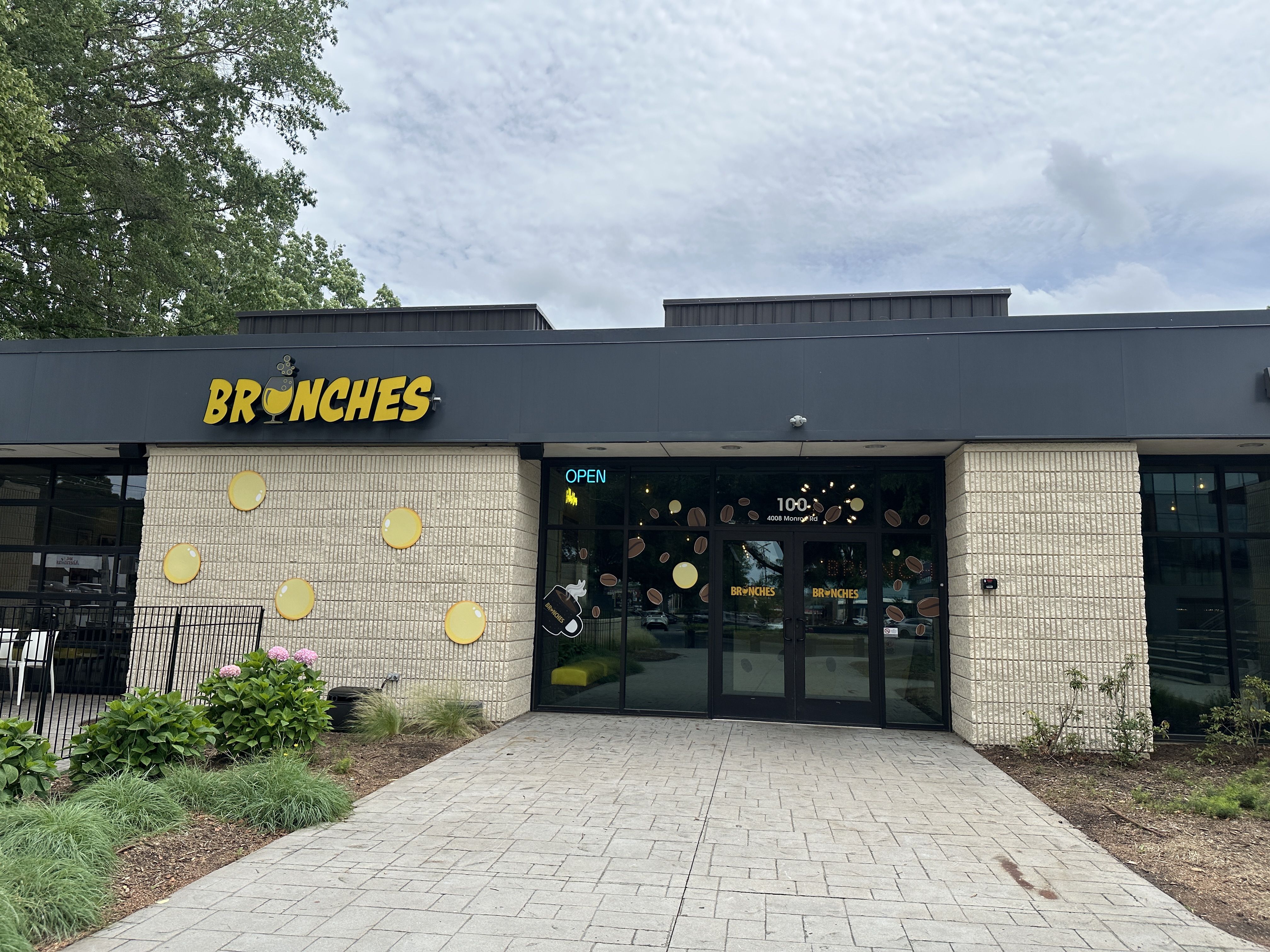 Storefront with a dark gray facade and a bright yellow sign reading BRONCHES with a wine glass icon. Wall decorated with large yellow discs; glass doors, open sign, greenery, and a cloudy sky.