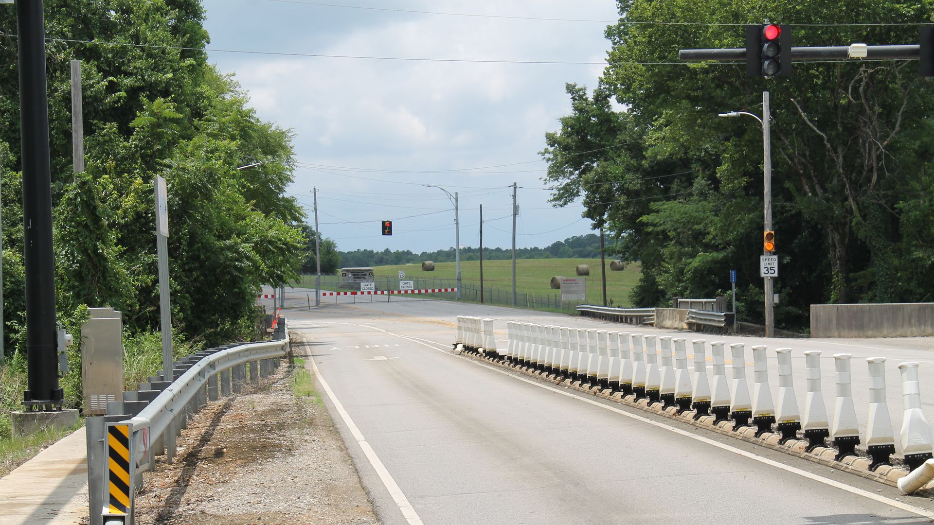 Rural road blocked by barricades and a red traffic light; white barrier pylons line the right edge while guard rails run along the road. Green trees flank fields with hay bales in the distance.